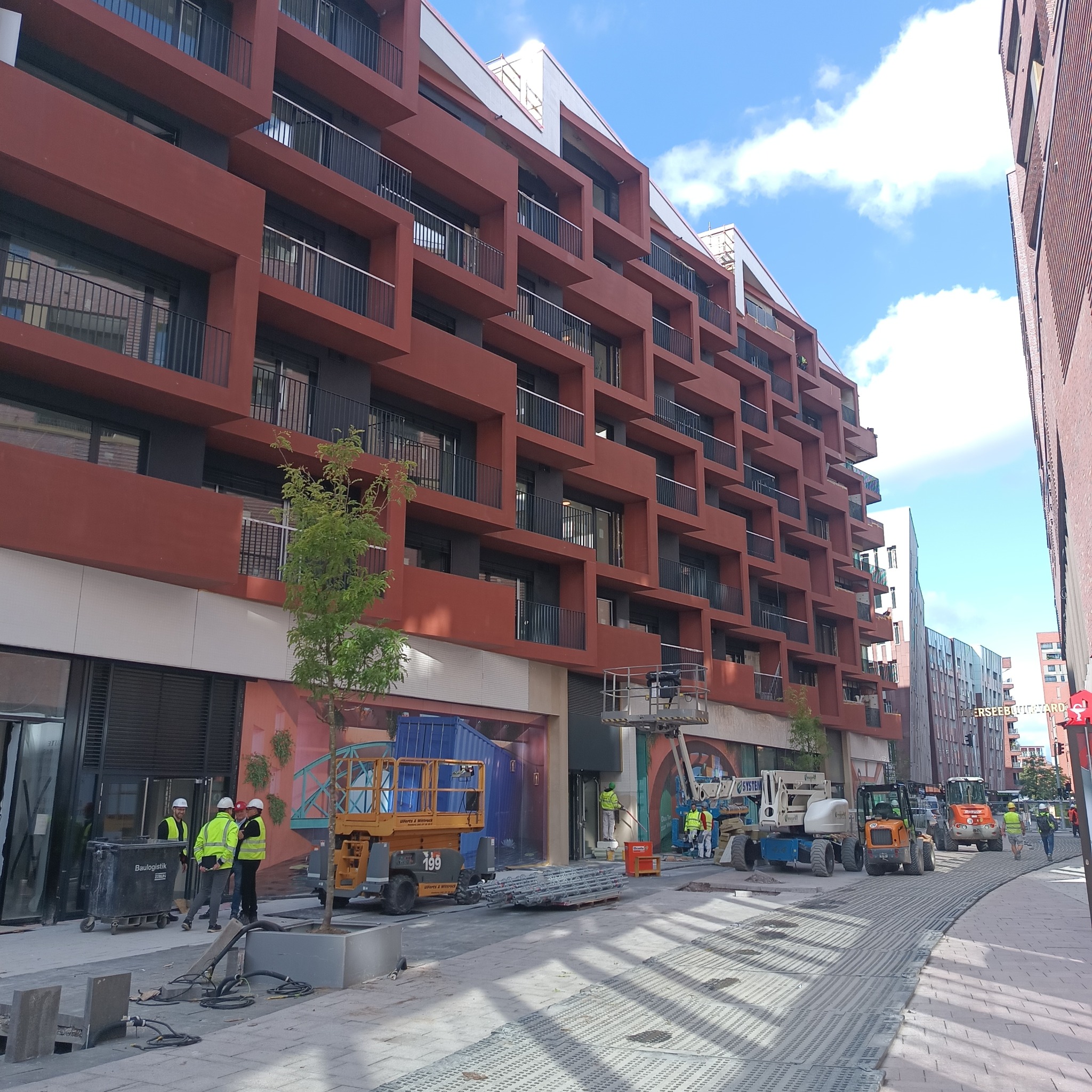 Prace instalacyjne w Nowej Wsi na zewnątrz nowoczesnego budynku z balkonami; widoczne podnośniki koszowe, robotnicy w kaskach i kamizelkach odblaskowych oraz przewody elektryczne.