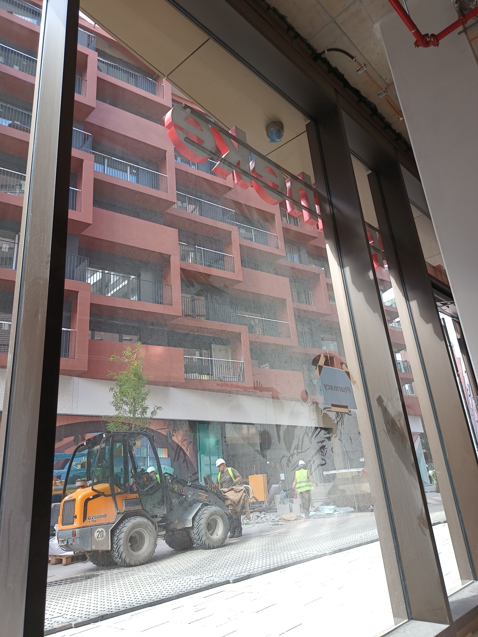 Widok przez okno na plac budowy z pracującymi robotnikami w kaskach i kamizelkach odblaskowych, używającymi małej ładowarki na tle budynku z balkonami i czerwonymi napisami.