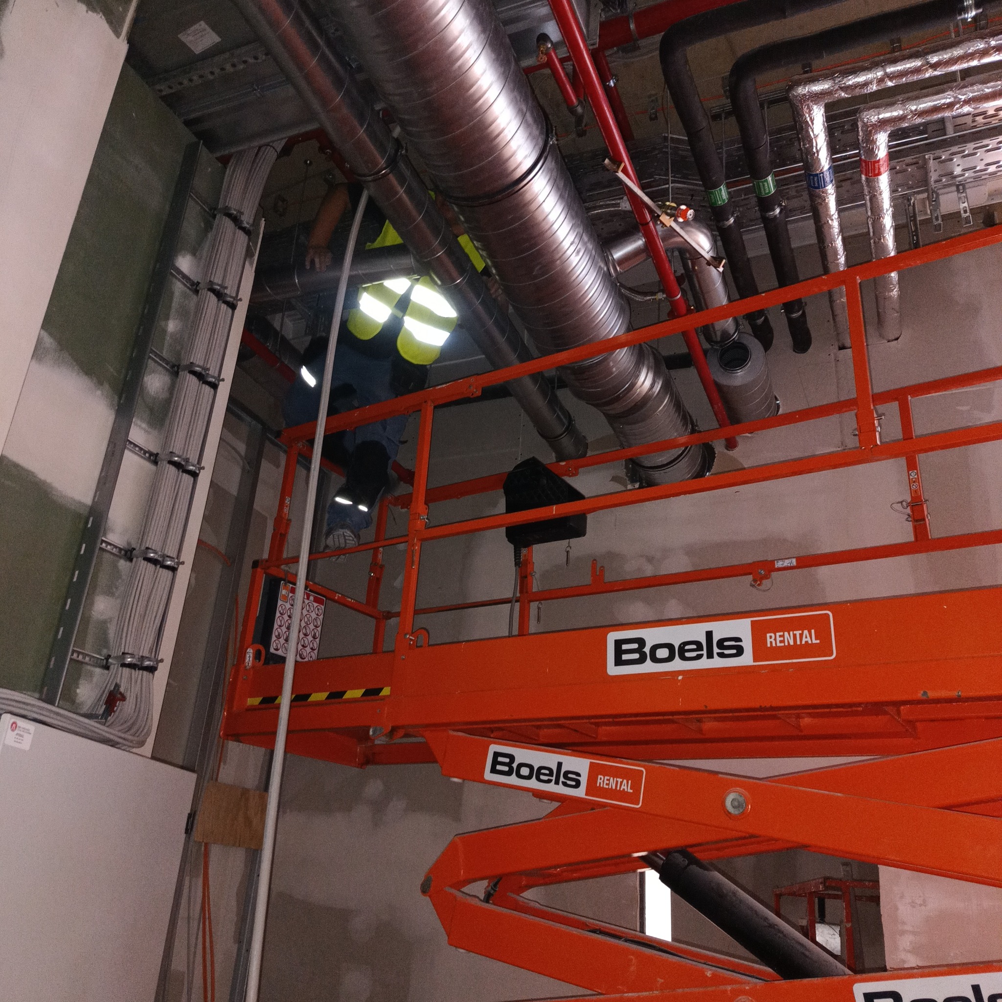 Prace instalacyjne w surowym wnętrzu: robotnik w kamizelce odblaskowej na podnośniku Boels Rental, prowadzi kable wzdłuż ściany, widoczne rury wentylacyjne pod sufitem.