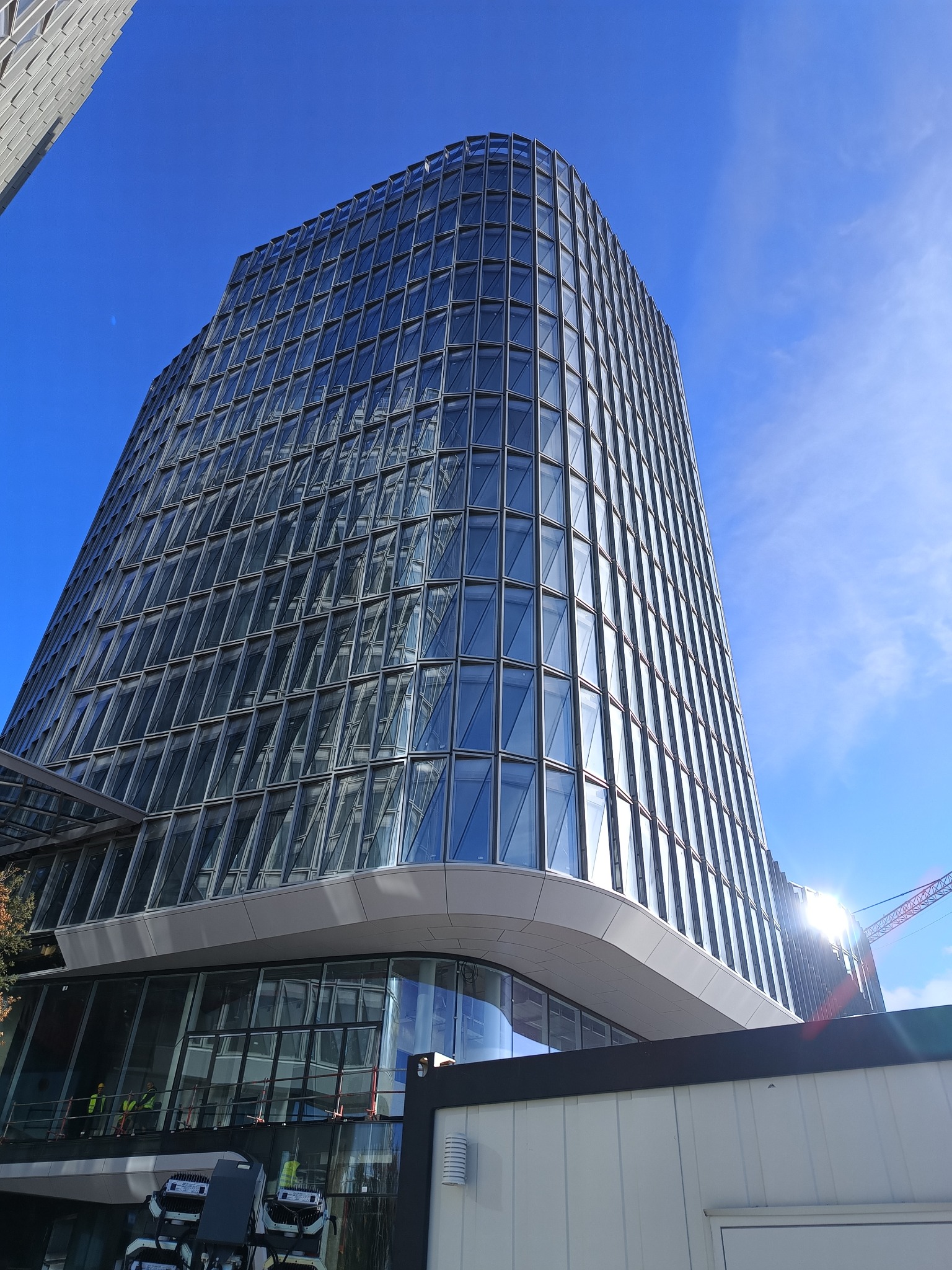 Nowoczesny budynek biurowy z przeszkloną fasadą i widocznymi pracownikami w żółtych kamizelkach odblaskowych na niższych poziomach, sugerującymi prace instalacyjne lub wykończeniowe, z jasnym...