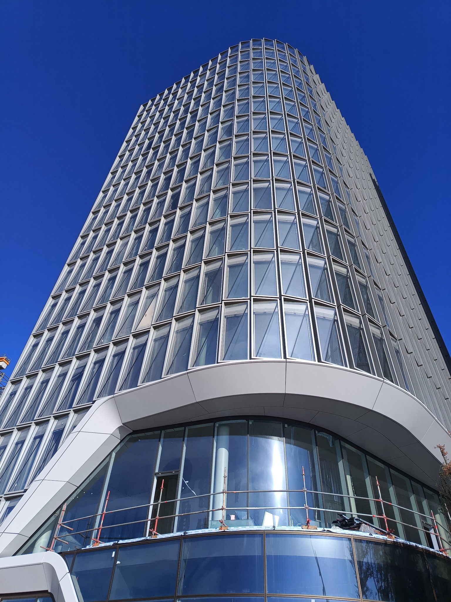 Wysoki, nowoczesny budynek biurowy z licznymi oknami, sfotografowany pod ostrym kątem z dołu, z widocznymi rusztowaniami na niższych kondygnacjach.