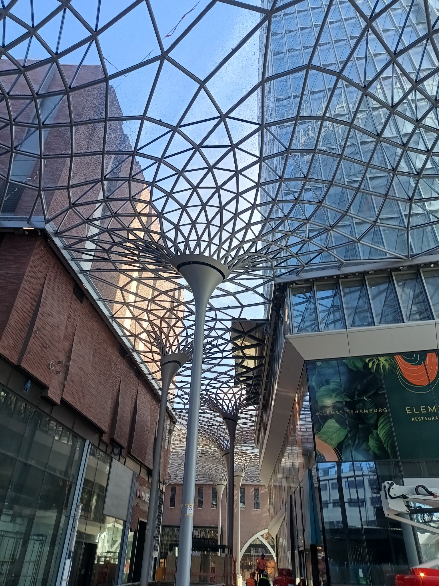 Szklany dach o konstrukcji kratownicowej z elementami oświetleniowymi w kształcie drzew, widok z dołu na budynki z cegły i szkła w tle, w tle widoczne prace budowlane.