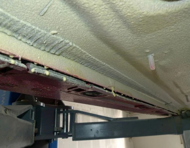 Spód samochodu na podnośniku po nałożeniu warstwy ochronnej, widoczne ślady spływającej substancji i fragment podnośnika.