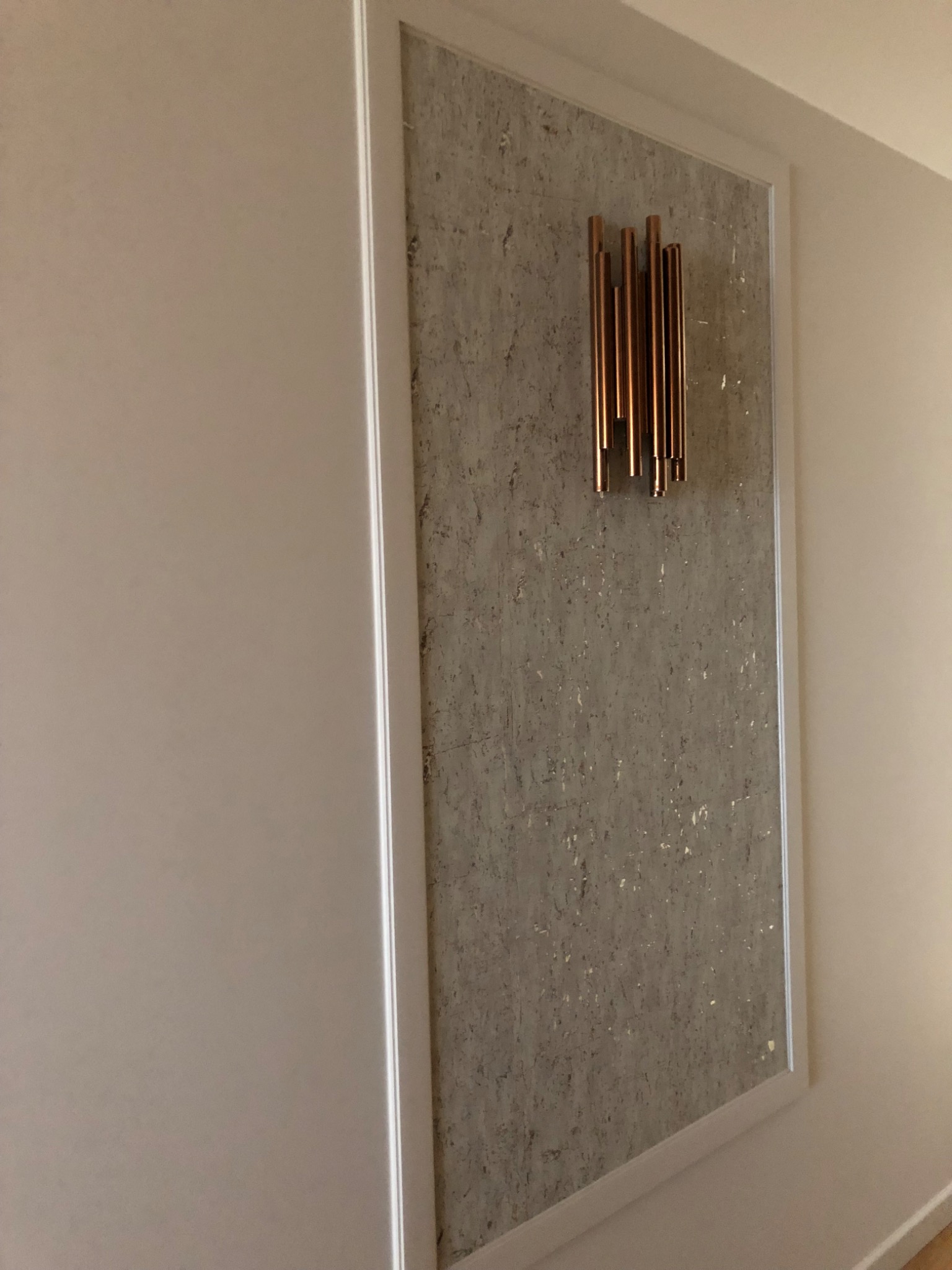 Dekoracyjny panel ścienny z szarego korka w białej ramie, ozdobiony pionowymi, miedzianymi rurkami o zróżnicowanej długości.
