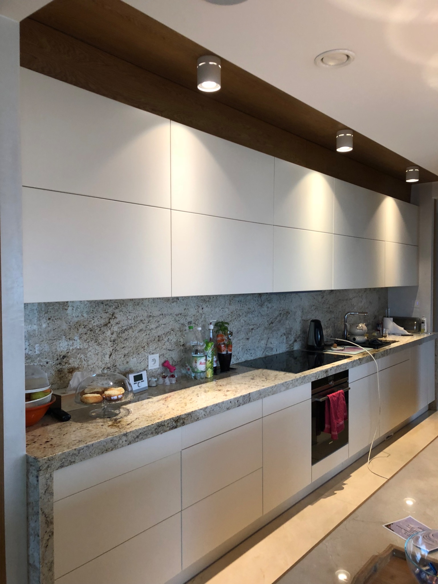 Nowoczesna kuchnia z białymi szafkami, blatem z jasnego granitu i pasującym backsplashiem, oświetlona punktowymi lampami pod drewnianą listwą sufitową, z widocznymi akcesoriami na blacie.