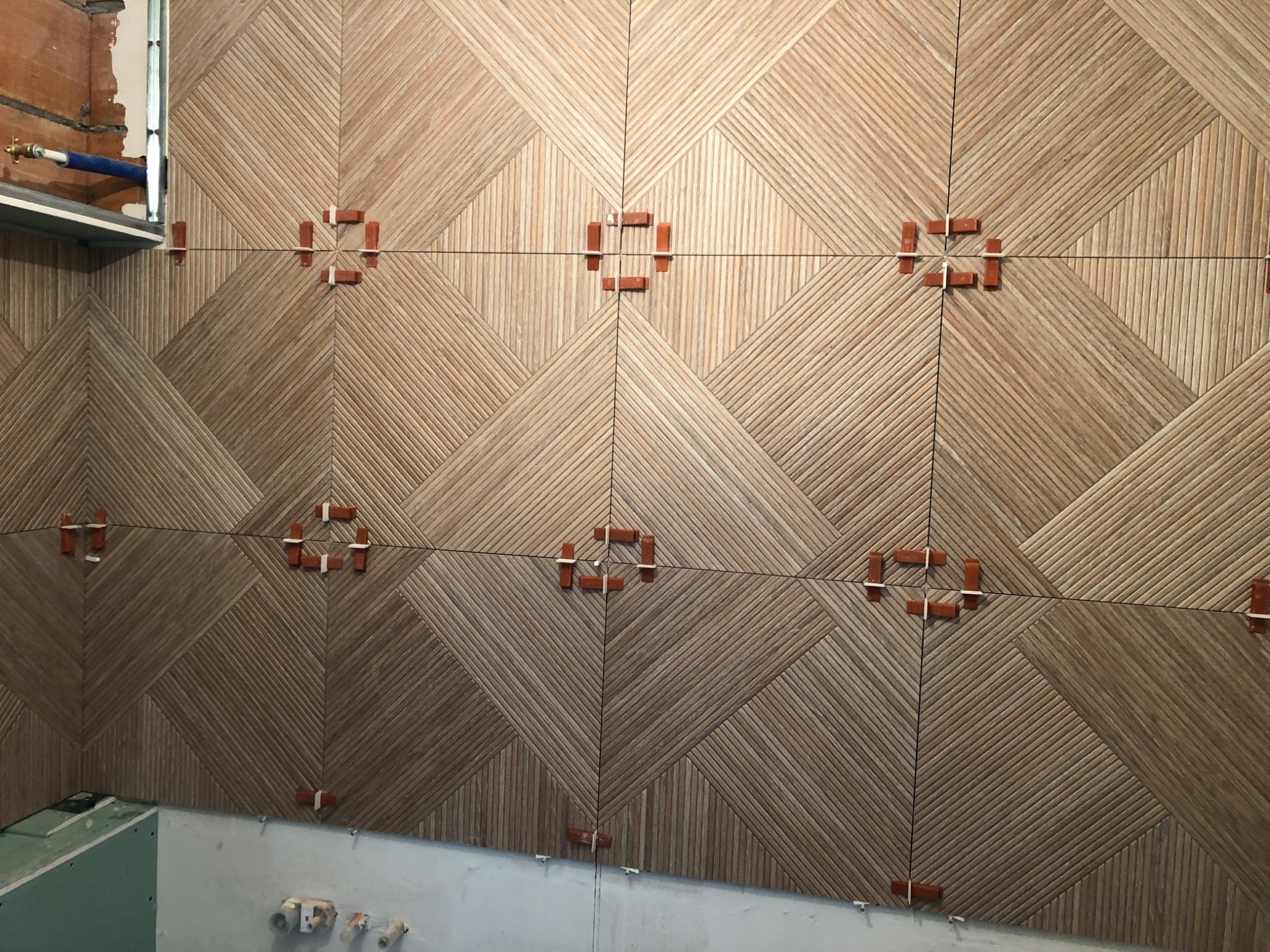 Ściana wyłożona płytkami imitującymi drewno, ułożonymi w geometryczny wzór z użyciem pomarańczowych krzyżyków dystansowych.