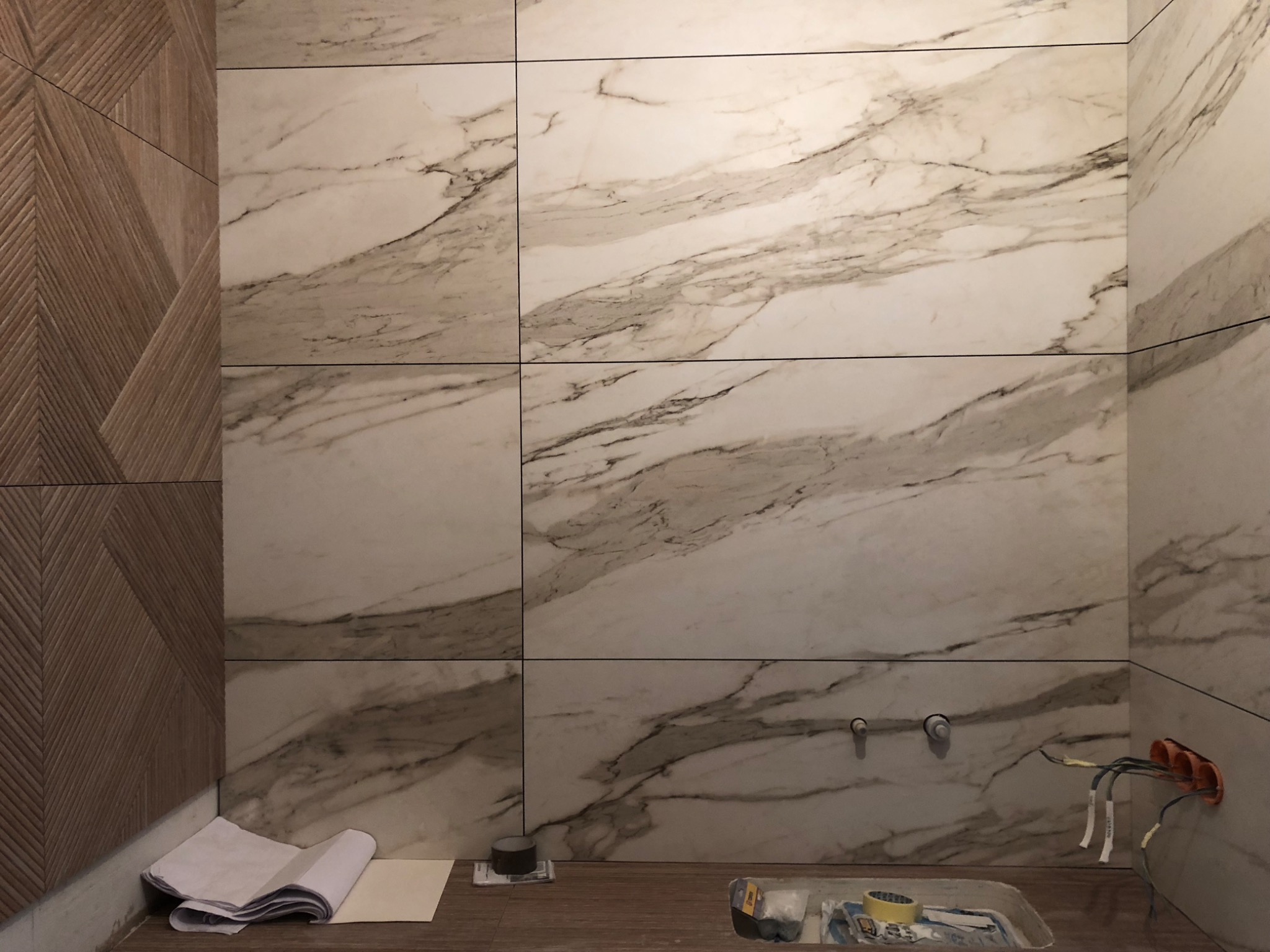 Łazienka w trakcie wykańczania: ściany wyłożone wielkoformatowymi płytkami imitującymi marmur, obok ściana z drewnopodobnych paneli z geometrycznym wzorem, widoczne wystające kable elektryczne...