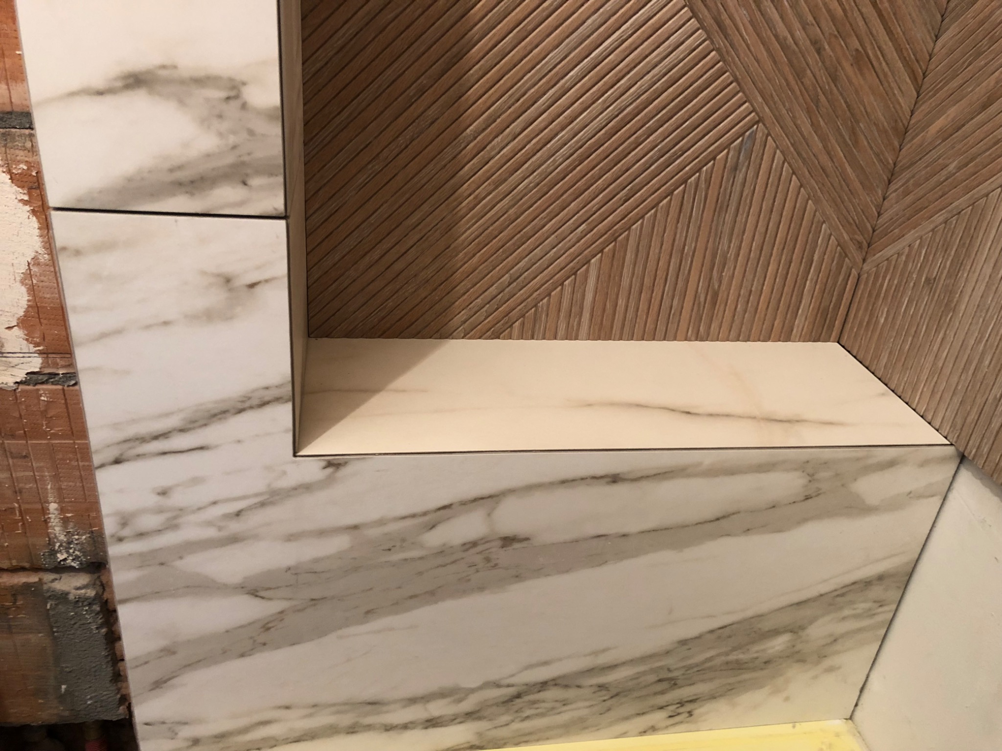 Wnęka w łazience wyłożona płytkami imitującymi marmur, z drewnopodobną ścianą z ukośnymi żłobieniami w tle, w trakcie montażu.