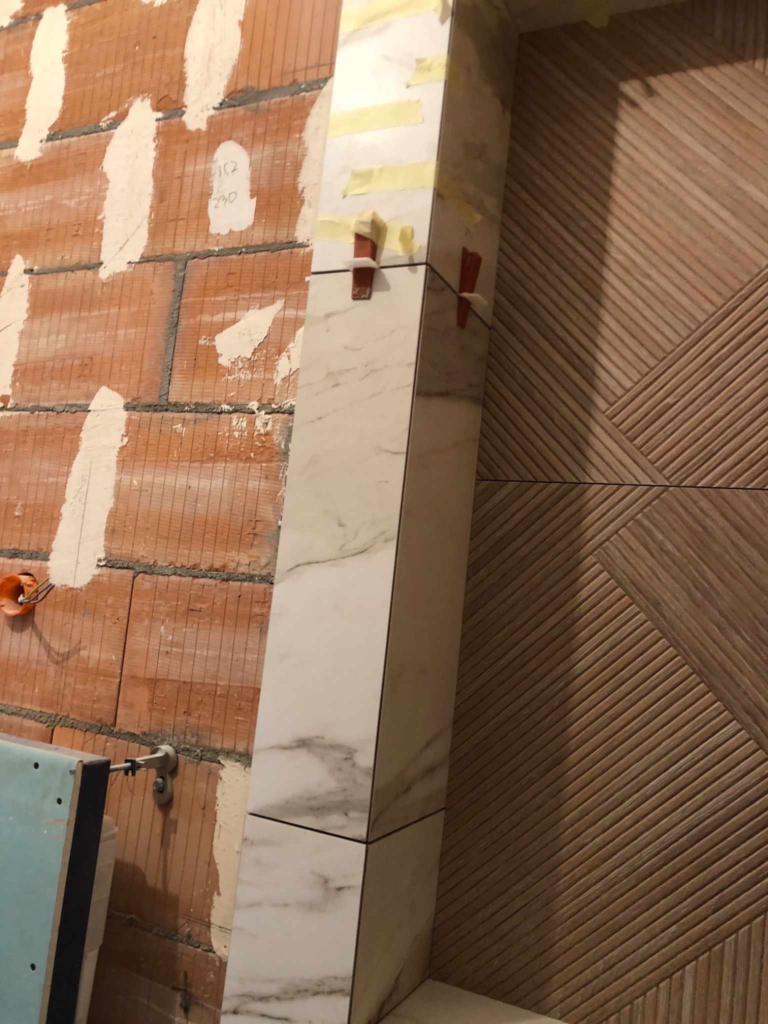 Montaż marmurowej ramy drzwiowej z użyciem klinów poziomujących, widoczna surowa ceglana ściana z instalacją elektryczną w tle oraz drewniana okładzina drzwi z geometrycznym wzorem.