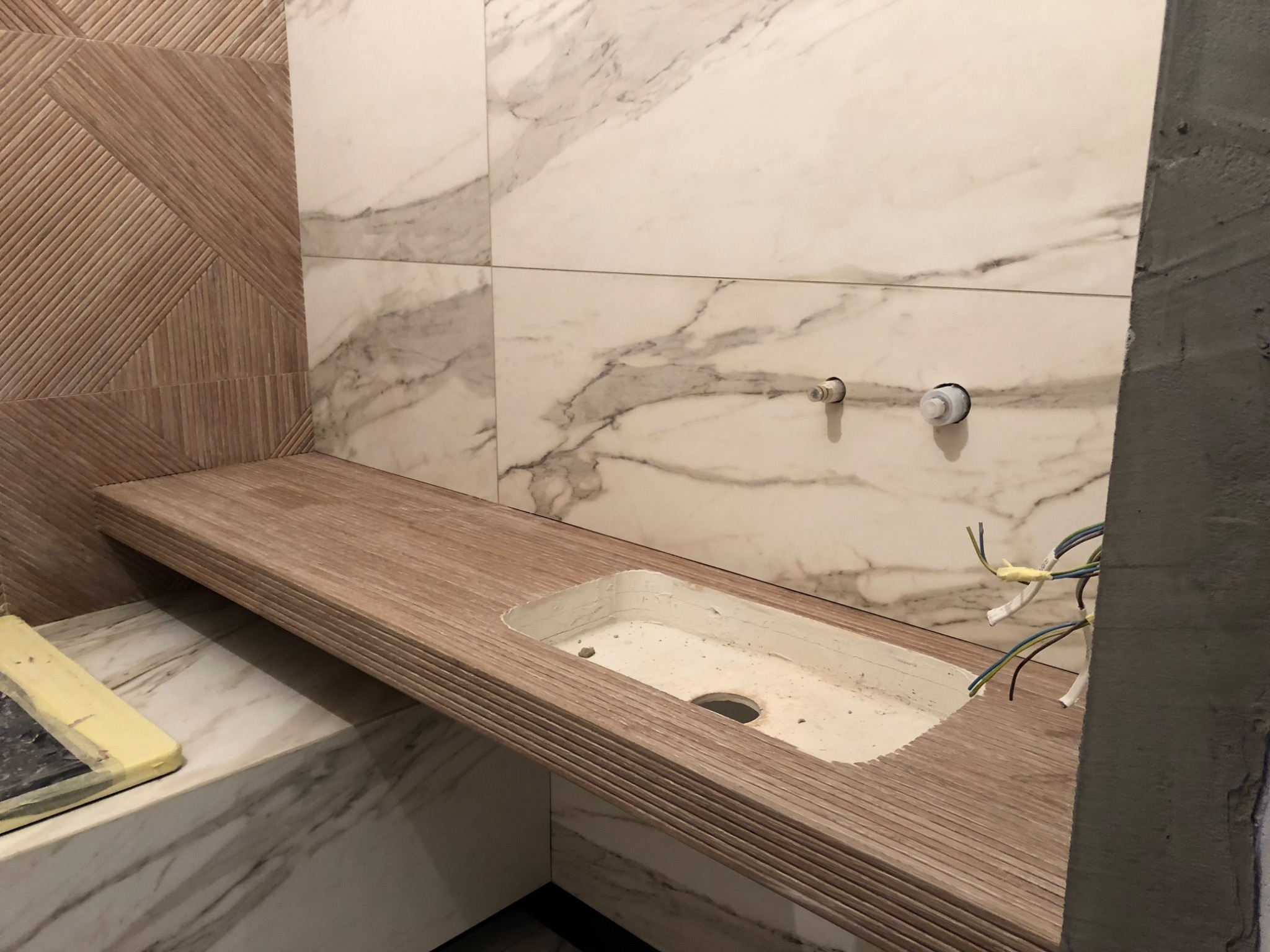 Łazienka w trakcie montażu blatu umywalkowego z frezowanymi krawędziami, widoczne przygotowane przyłącza hydrauliczne i elektryczne, ściany wyłożone płytkami imitującymi marmur i drewno.