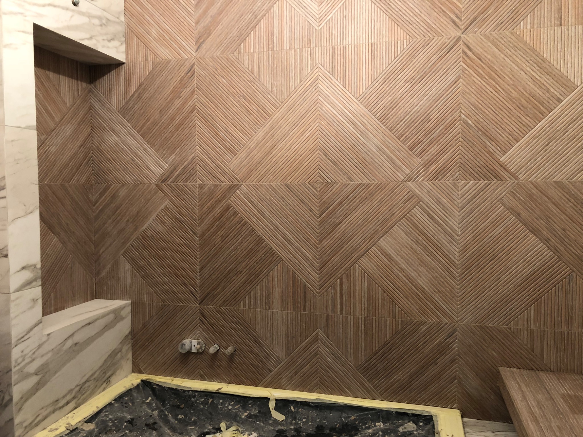 Łazienka w trakcie remontu: ściana wyłożona brązowymi płytkami z geometrycznym wzorem w jodełkę, marmurowa wnęka prysznicowa, widoczne elementy instalacji wodnej i zabezpieczona folią podłoga.