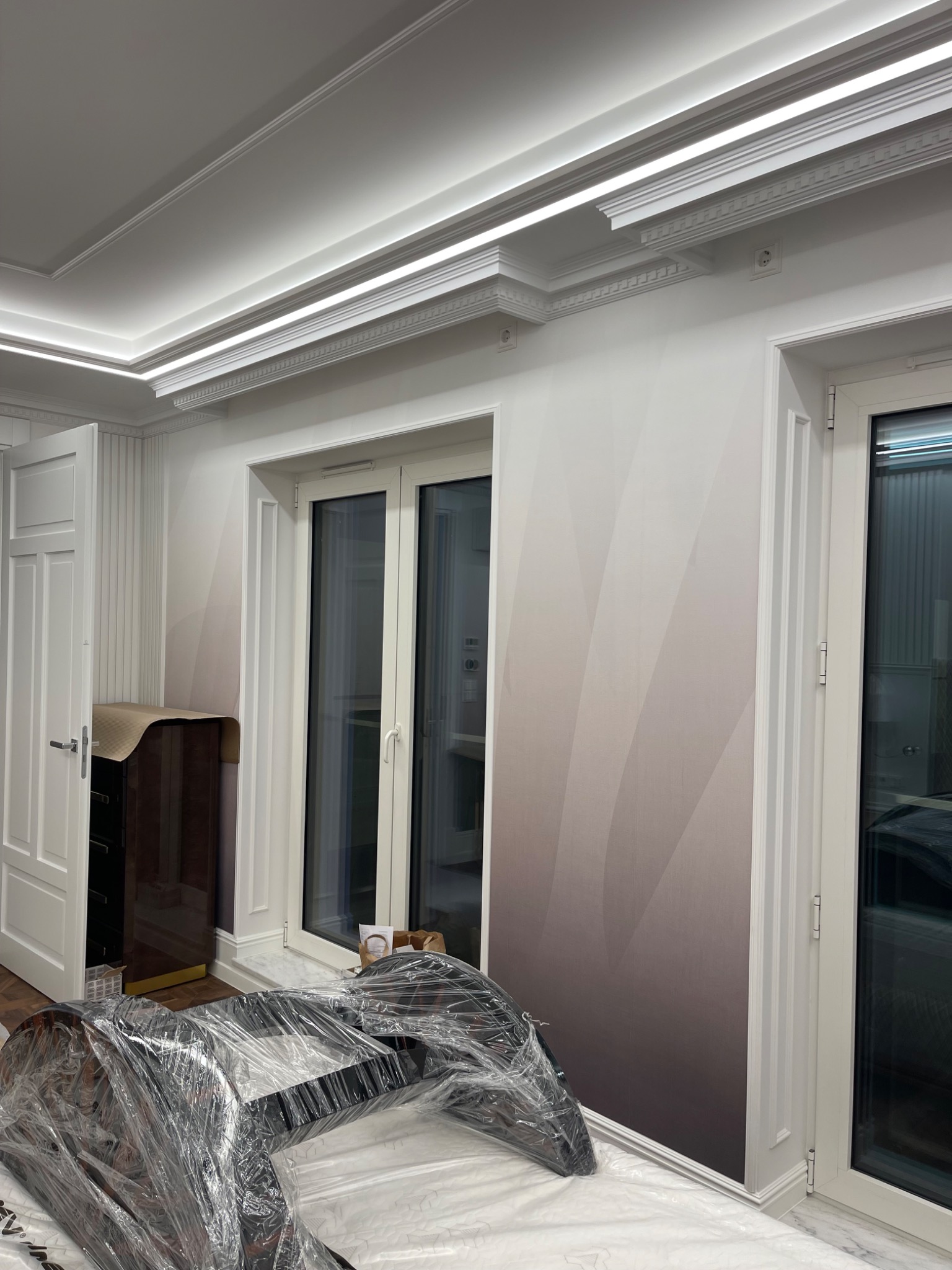 Pokój po remoncie z dekoracyjnymi listwami sufitowymi z oświetleniem LED, gradientową tapetą w odcieniach szarości i białymi drzwiami oraz oknami. Mebel zabezpieczony folią ochronną.