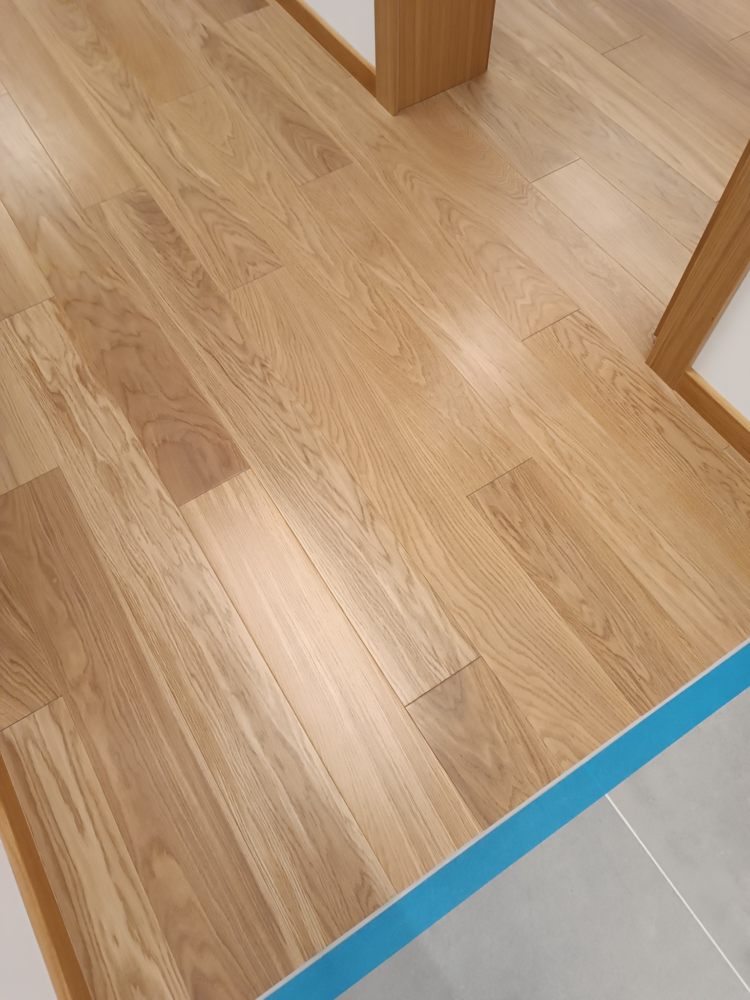 Jasne, drewniane deski podłogowe ułożone równolegle, widoczne przy listwie przypodłogowej i przejściu do płytek. Zabezpieczone niebieską taśmą malarską.