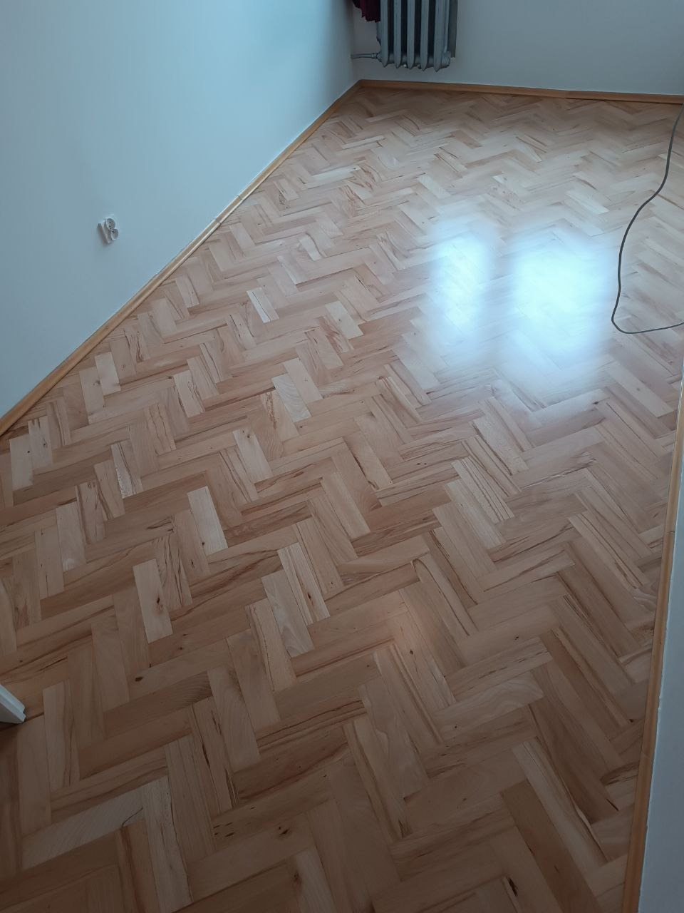 Ułożony parkiet w jodełkę z jasnego drewna, widoczny fragment grzejnika i ściany z gniazdkiem.