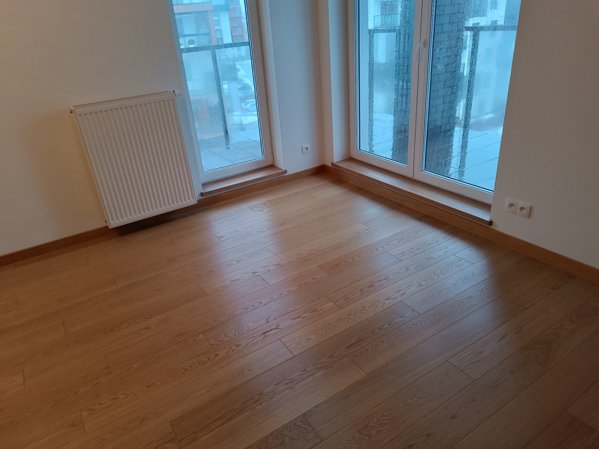 Jasny parkiet ułożony we wzór prosty w pustym pokoju z dwoma oknami balkonowymi i grzejnikiem na ścianie.