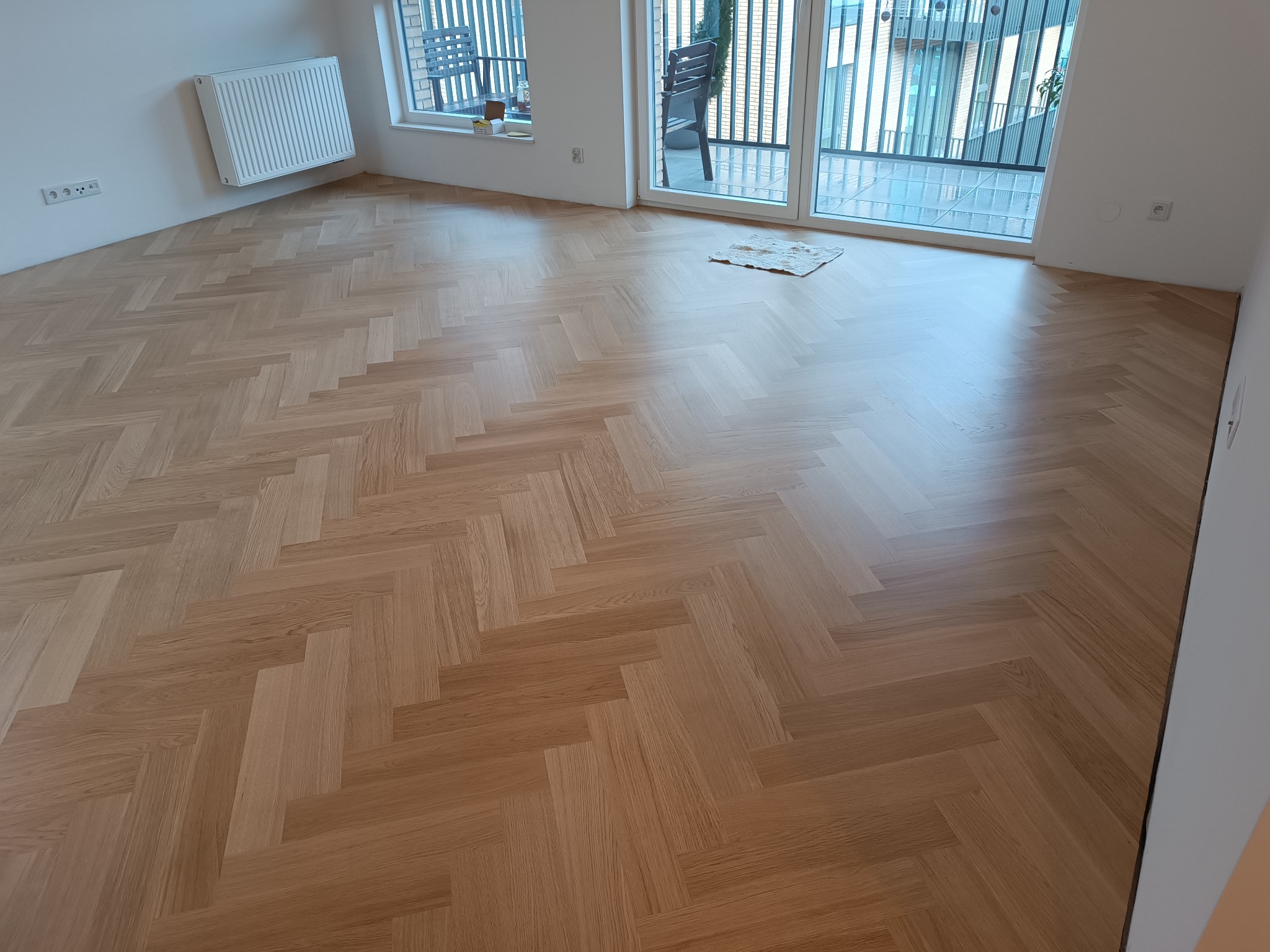 Ułożony parkiet w jodełkę, jasny, lakierowany, widok na balkon z krzesłami, białe ściany, grzejnik