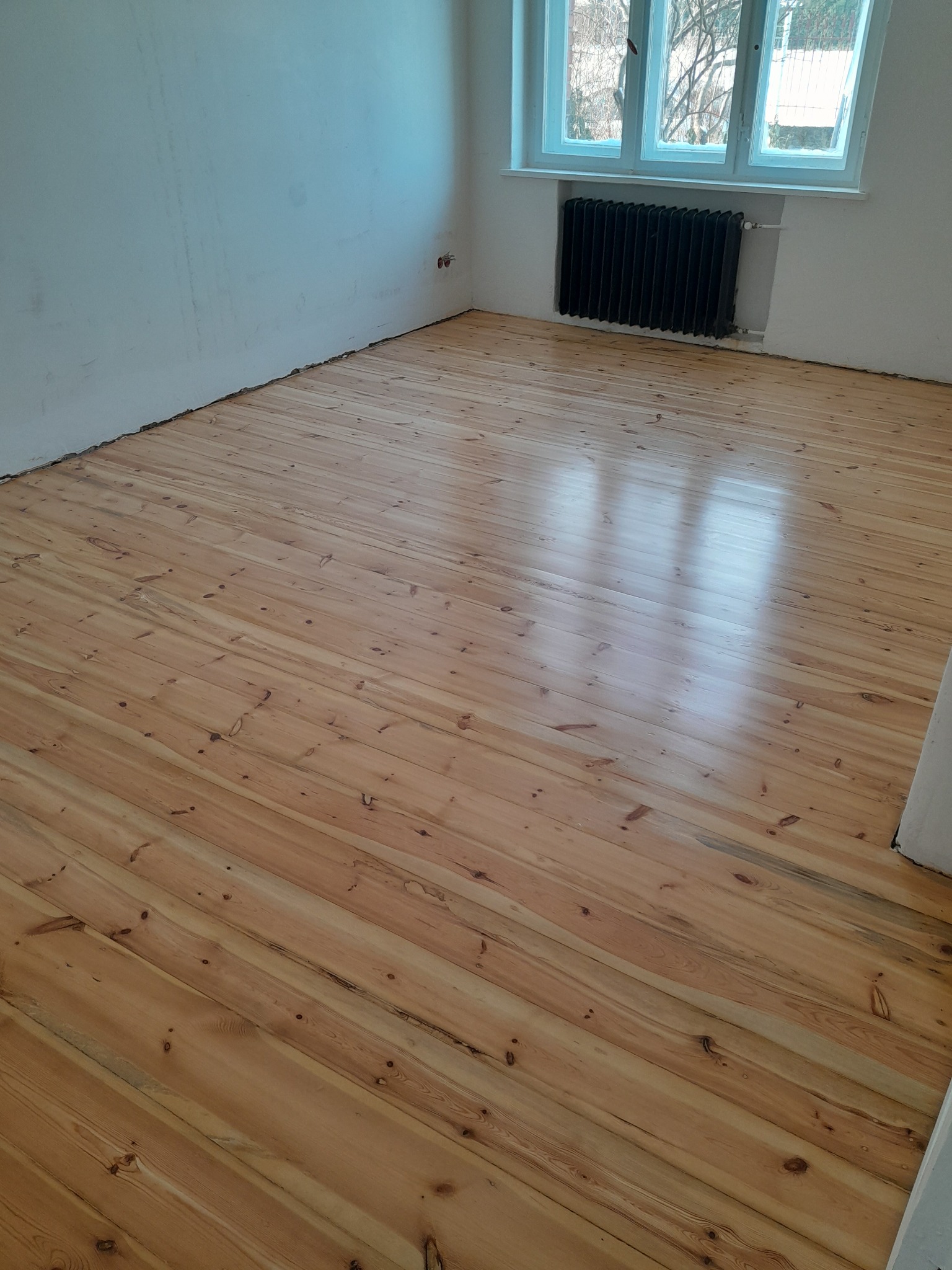 Jasna, odnowiona podłoga z desek w pokoju z grzejnikiem pod oknem. Ślady niedokładnego szpachlowania na ścianie.