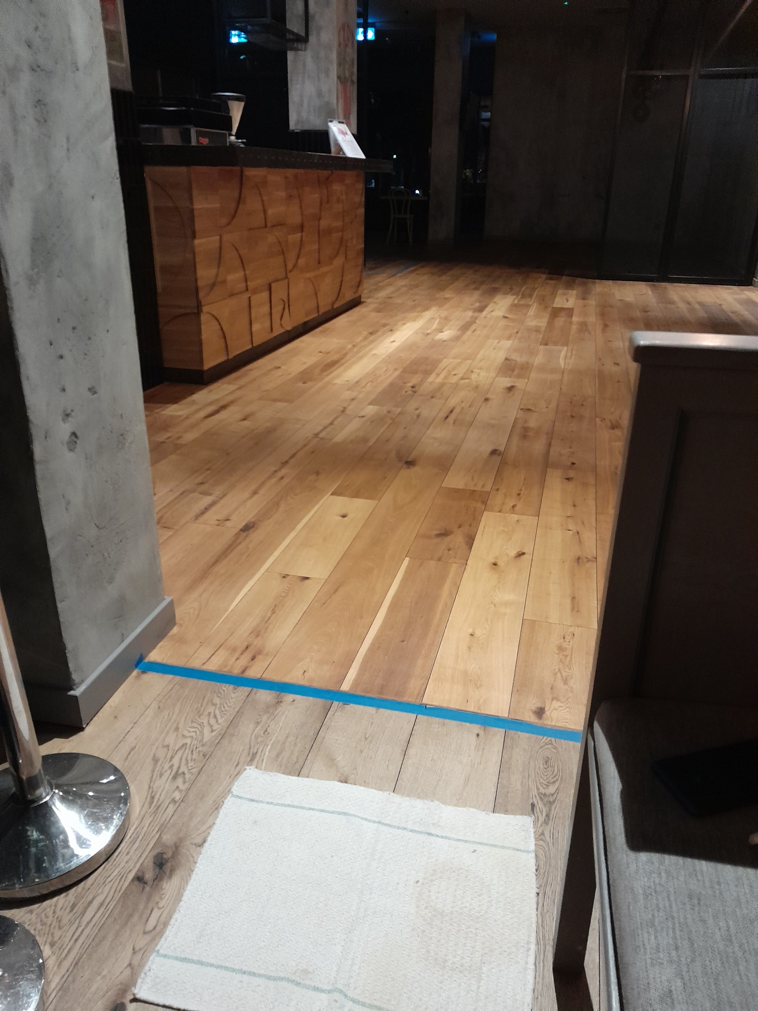 Jasne drewniane deski podłogowe z widocznym usłojeniem, oddzielone niebieską taśmą od ciemniejszej podłogi, z fragmentem dywanika i nogi mebla w kadrze.