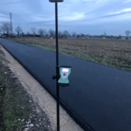 Geodezyjny odbiornik GPS RTK na tyczce, ustawiony przy nowo położonym asfalcie, w tle pole i słup telekomunikacyjny, pochmurne niebo.