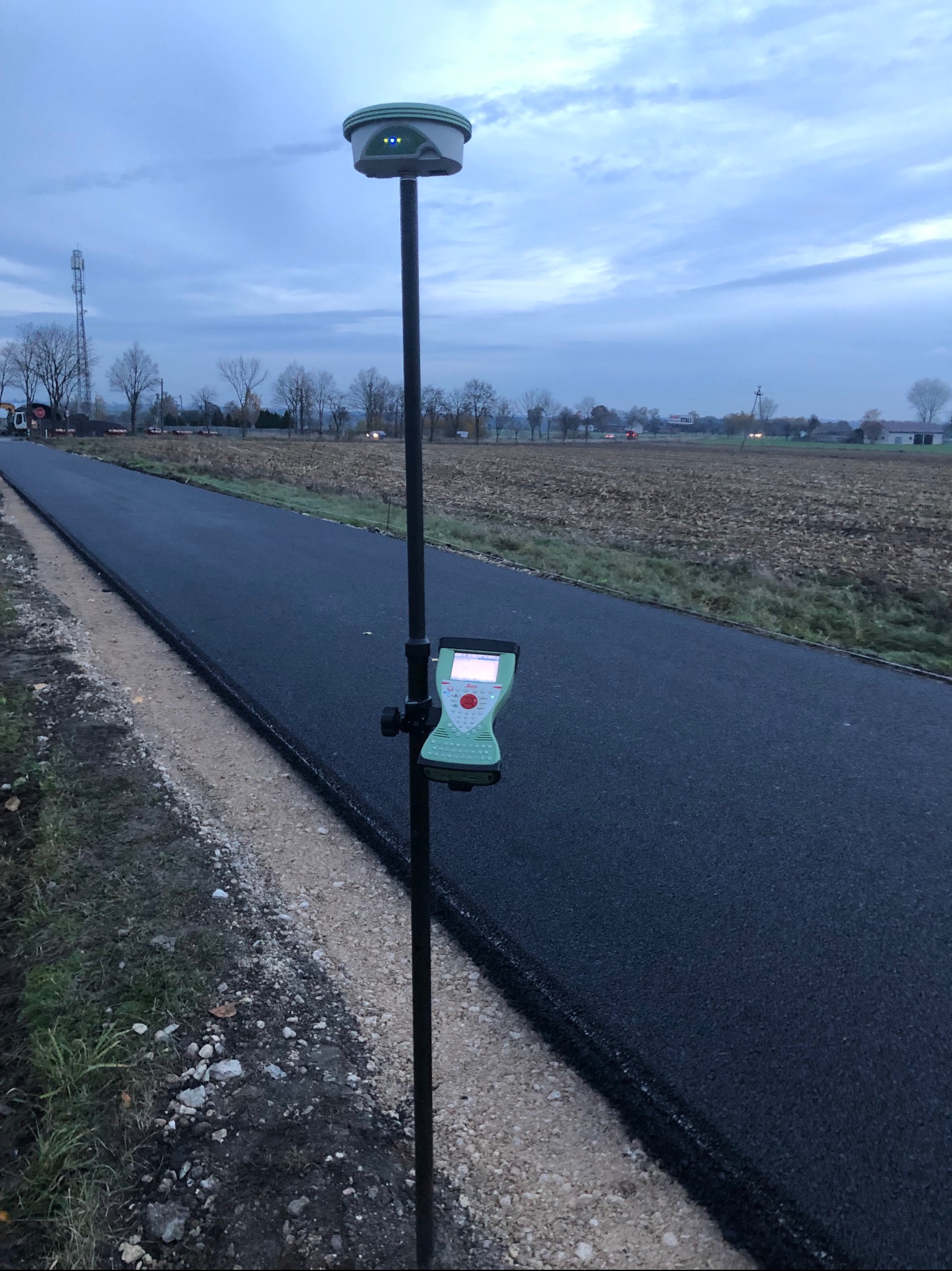 Geodezyjny odbiornik GPS RTK na tyczce, ustawiony przy nowo położonym asfalcie, w tle pole i słup telekomunikacyjny, pochmurne niebo.