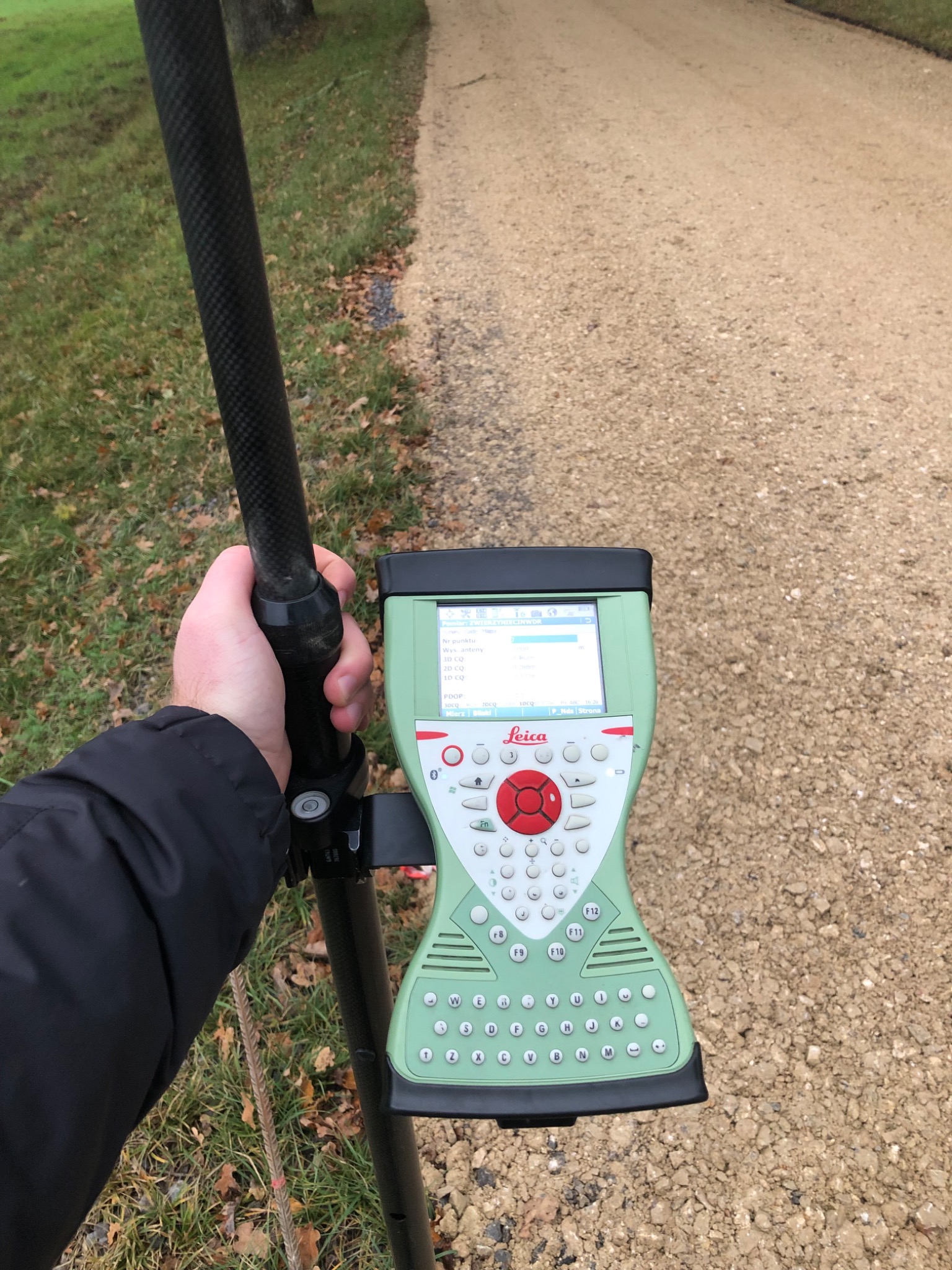 Pomiar geodezyjny w Bełchatowie: ręka trzyma tyczkę geodezyjną z zamontowanym na niej zielonym odbiornikiem GPS Leica, ustawioną na żwirowej drodze, w tle trawa i drzewa.