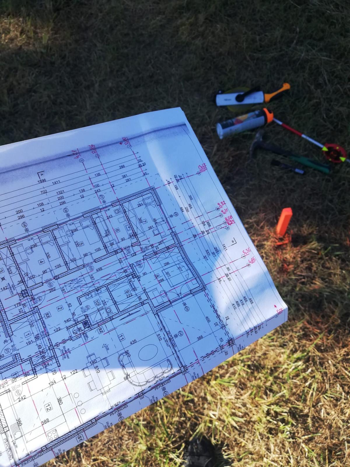 Plan architektoniczny budynku rozłożony na trawie obok narzędzi geodezyjnych i pomiarowych, takich jak dalmierz laserowy, spray znakujący i paliki. Widoczny fragment planu z detalami pomieszczeń...