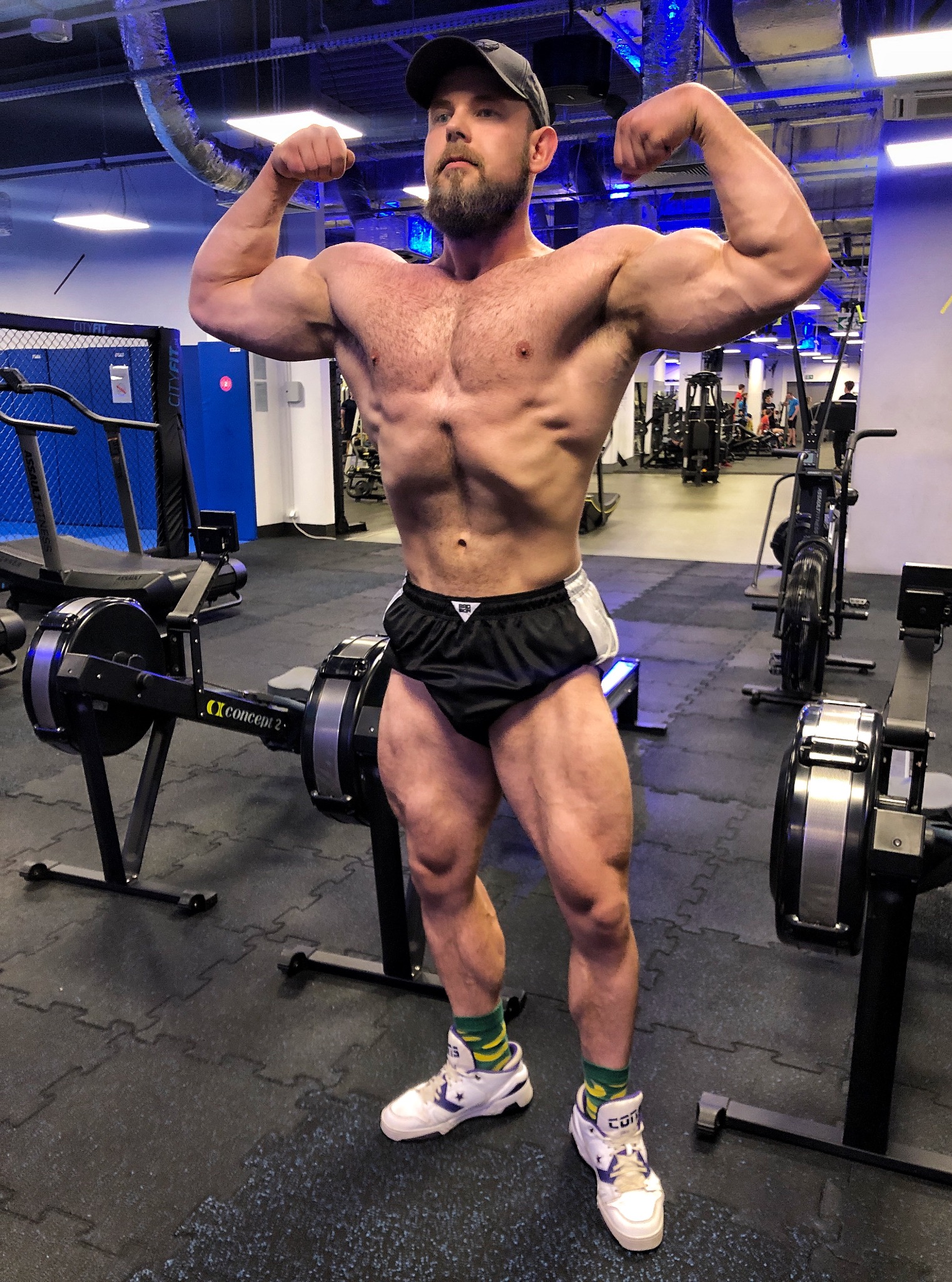 Mężczyzna w sportowym stroju prezentuje muskulaturę na tle siłowni z maszynami do ćwiczeń. Widoczne zarysowane mięśnie brzucha, bicepsów i ud. Uśmiech na twarzy.