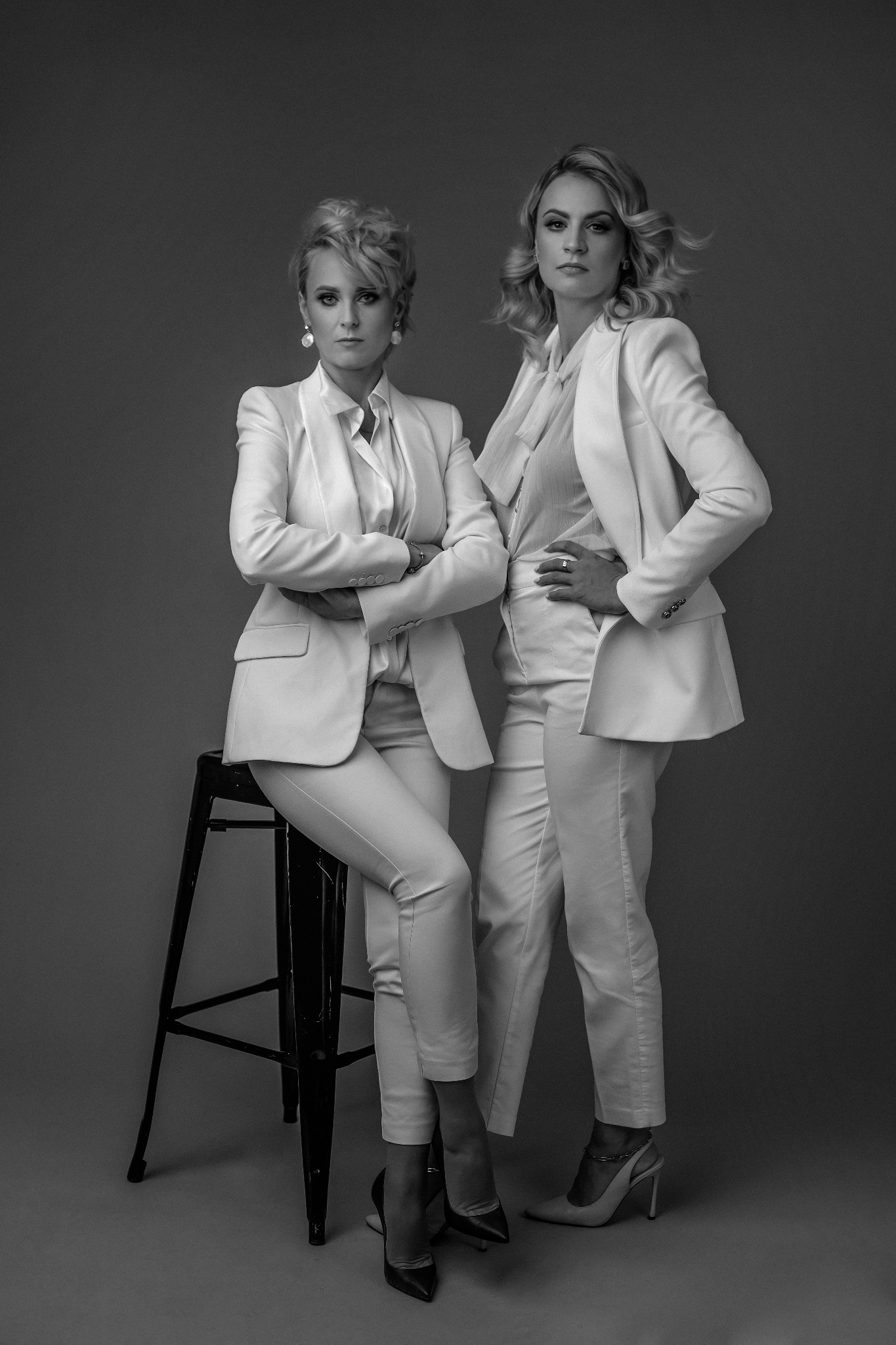 Dwie kobiety w białych garniturach pozują na szarym tle. Jedna siedzi na czarnym stołku barowym z założonymi rękami, druga stoi z ręką na biodrze. Zdjęcie czarno-białe, studyjne.
