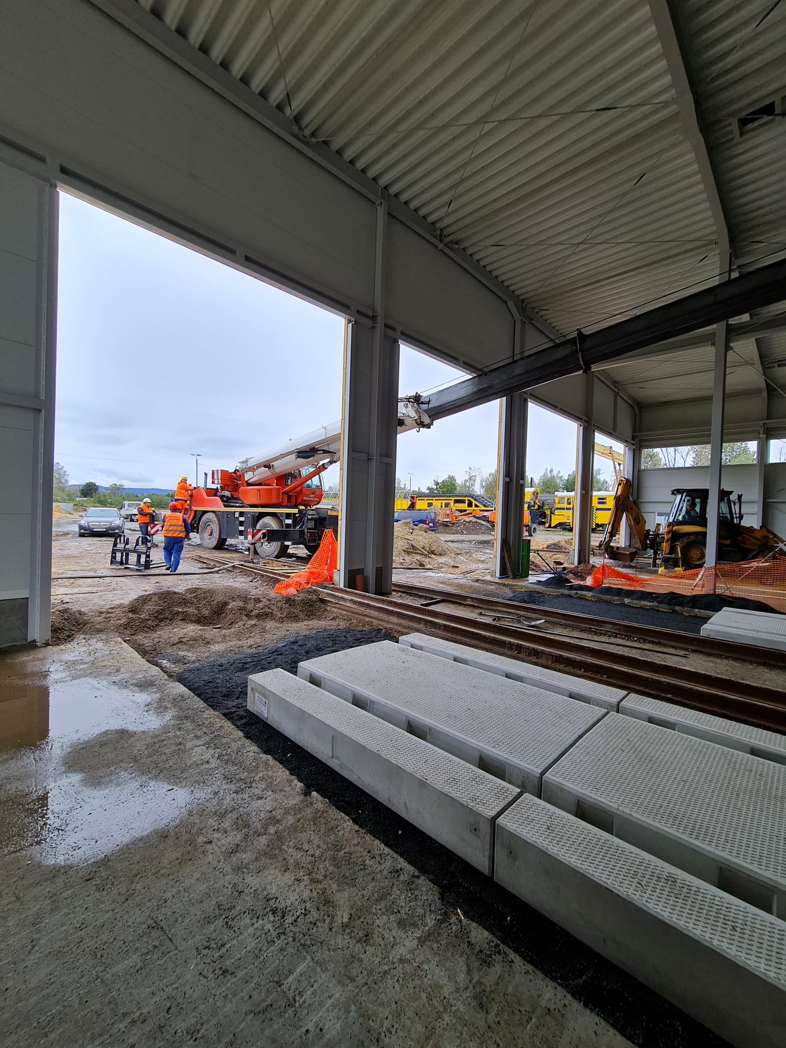 Plac budowy z żurawiem i koparką w hali, widoczne prefabrykaty betonowe, pracownicy w odblaskowych kamizelkach, teren po deszczu.