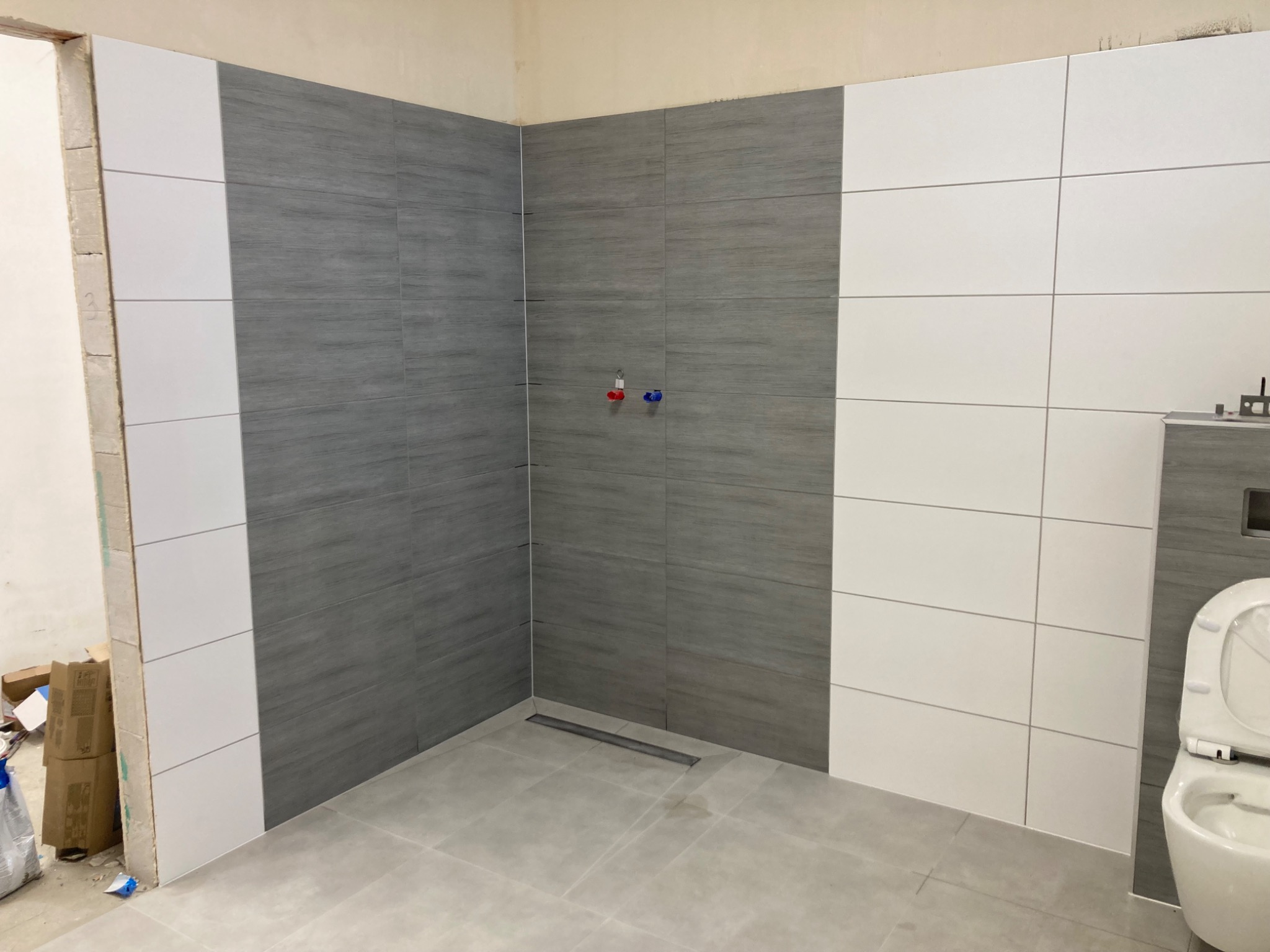 Wrocław: Wnętrze łazienki w trakcie układania płytek, widoczny fragment ściany z szarymi płytkami imitującymi drewno, białe płytki po bokach, odpływ liniowy w podłodze i częściowo zamontowana toaleta.