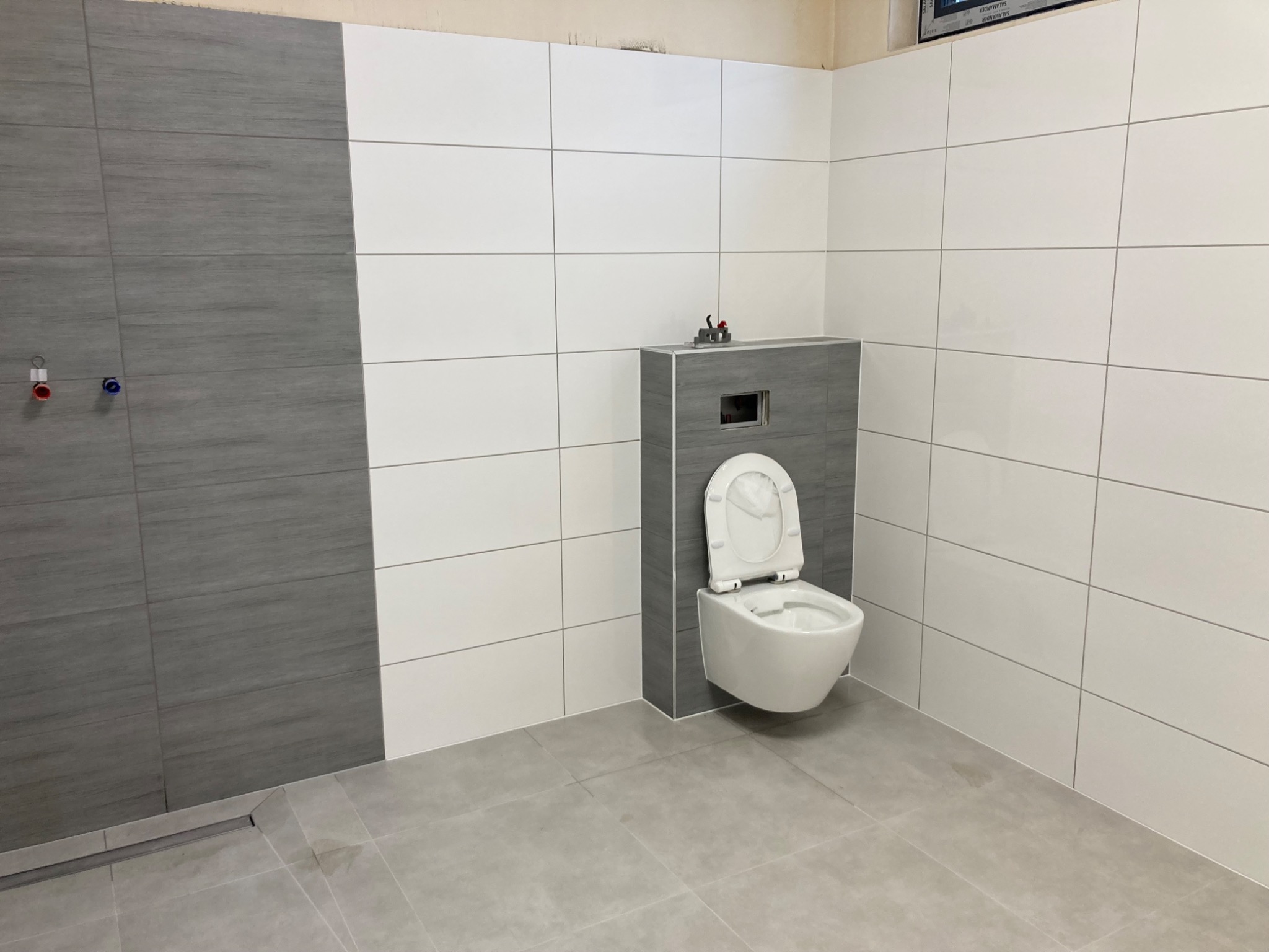 Nowoczesna toaleta podwieszana w szaro-białej łazience z odpływem liniowym w podłodze, widoczne przyłącza wodne.