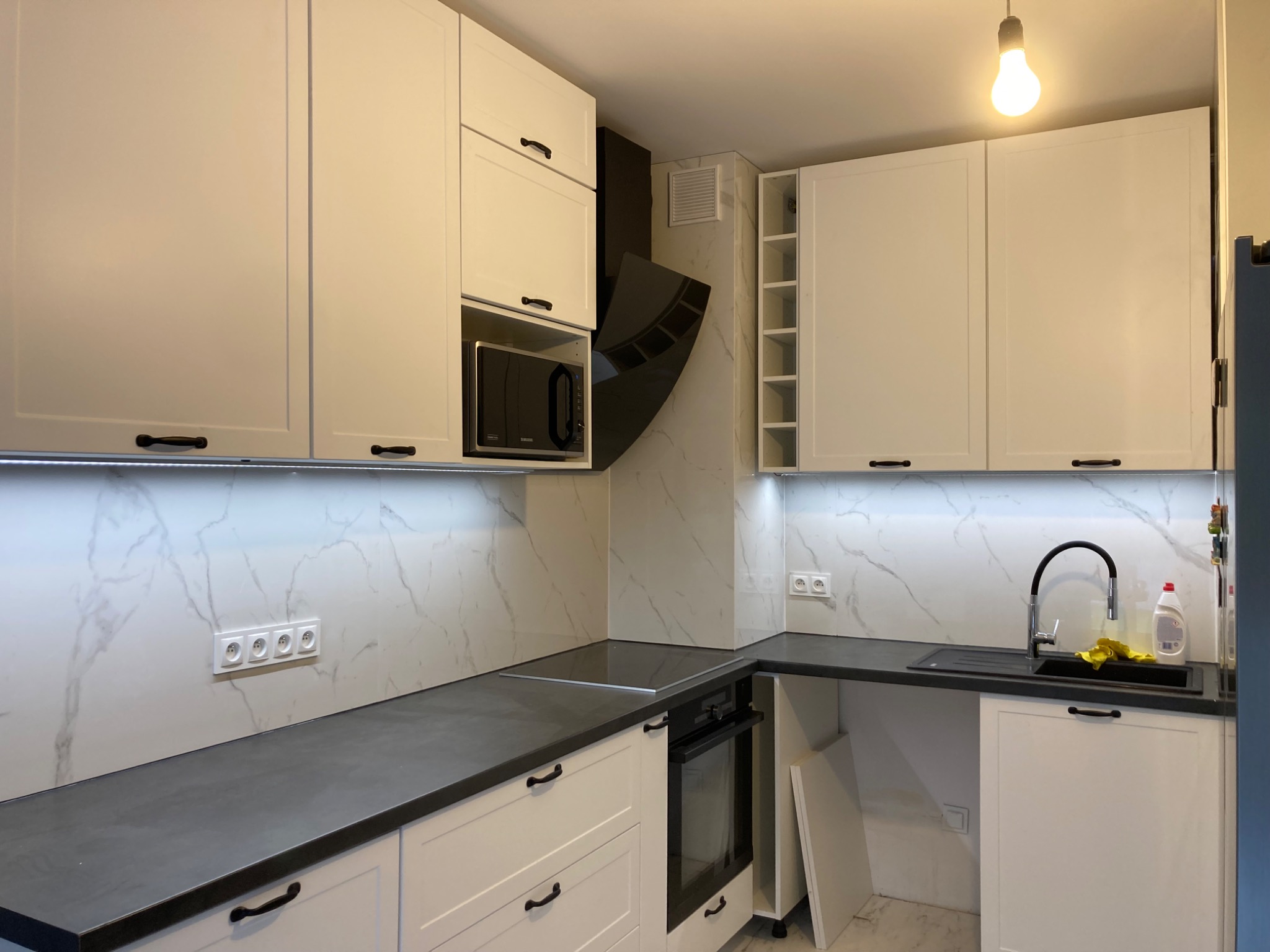 Nowoczesna kuchnia z białymi szafkami, czarnym blatem i marmurową ścianą, oświetlona listwą LED pod szafkami wiszącymi i pojedynczą żarówką.