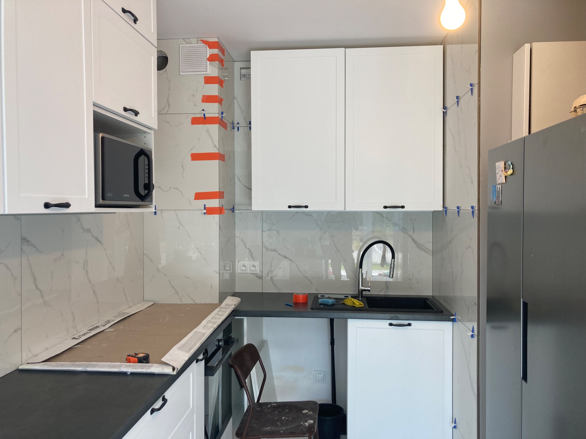 Kuchnia w trakcie wykańczania: białe szafki z czarnymi uchwytami, marmurowe płytki na ścianie z pomarańczową taśmą i niebieskimi krzyżykami dystansowymi, czarny zlew i blat, widoczny fragment...