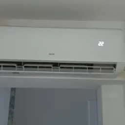 Biała klimatyzacja marki AUX zamontowana na ścianie, wyświetlacz pokazuje temperaturę 22 stopnie Celsjusza, widoczne niedoskonałości wykończenia ściany w rogu.