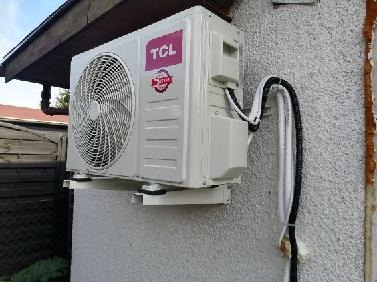 Zewnętrzna jednostka klimatyzacji TCL zamontowana na białym wsporniku na szarej, chropowatej ścianie budynku. Widoczne czarne i białe przewody prowadzące do jednostki.
