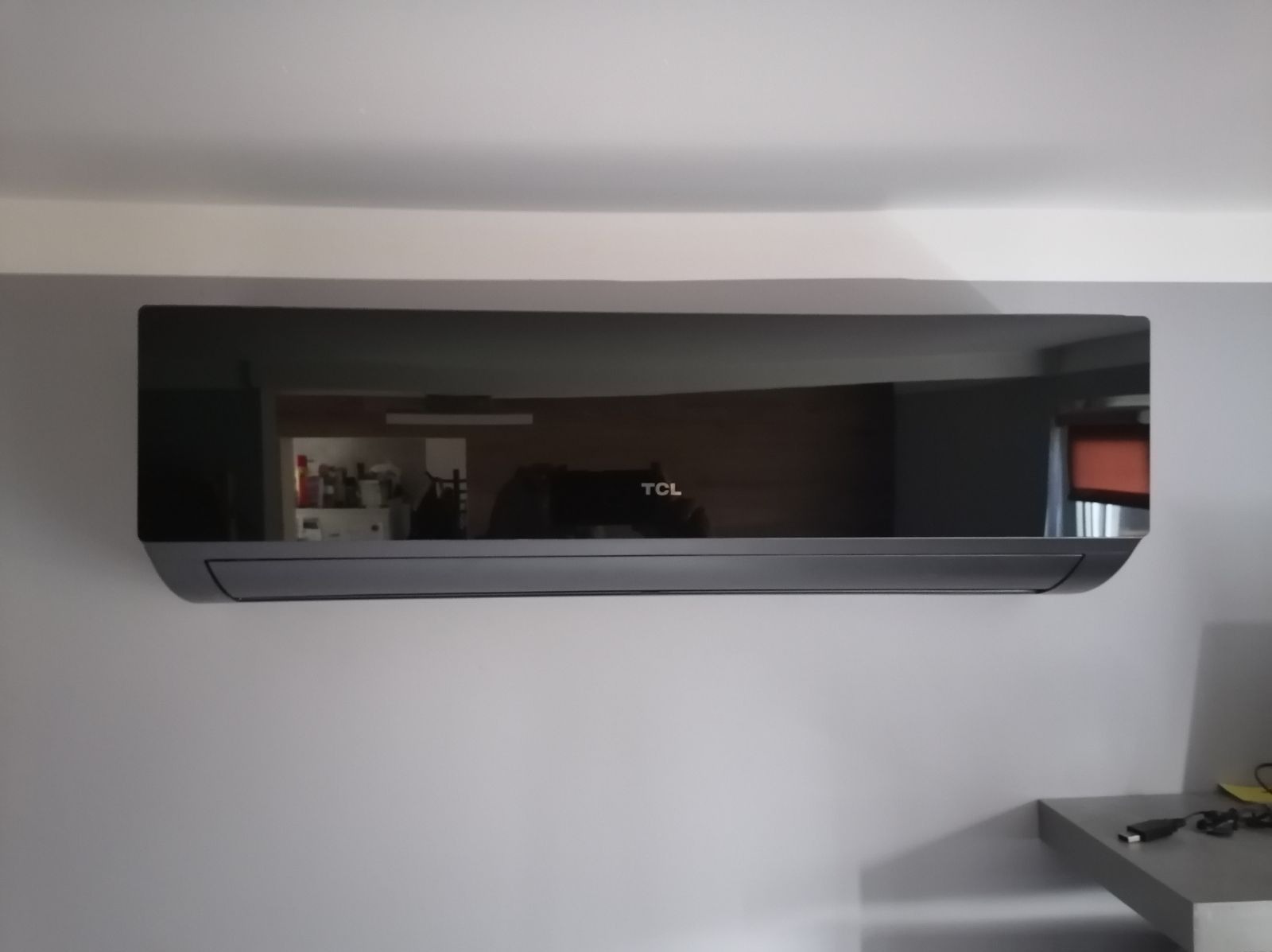 Czarny klimatyzator TCL zamontowany na białej ścianie, widoczne odbicie wnętrza pokoju.