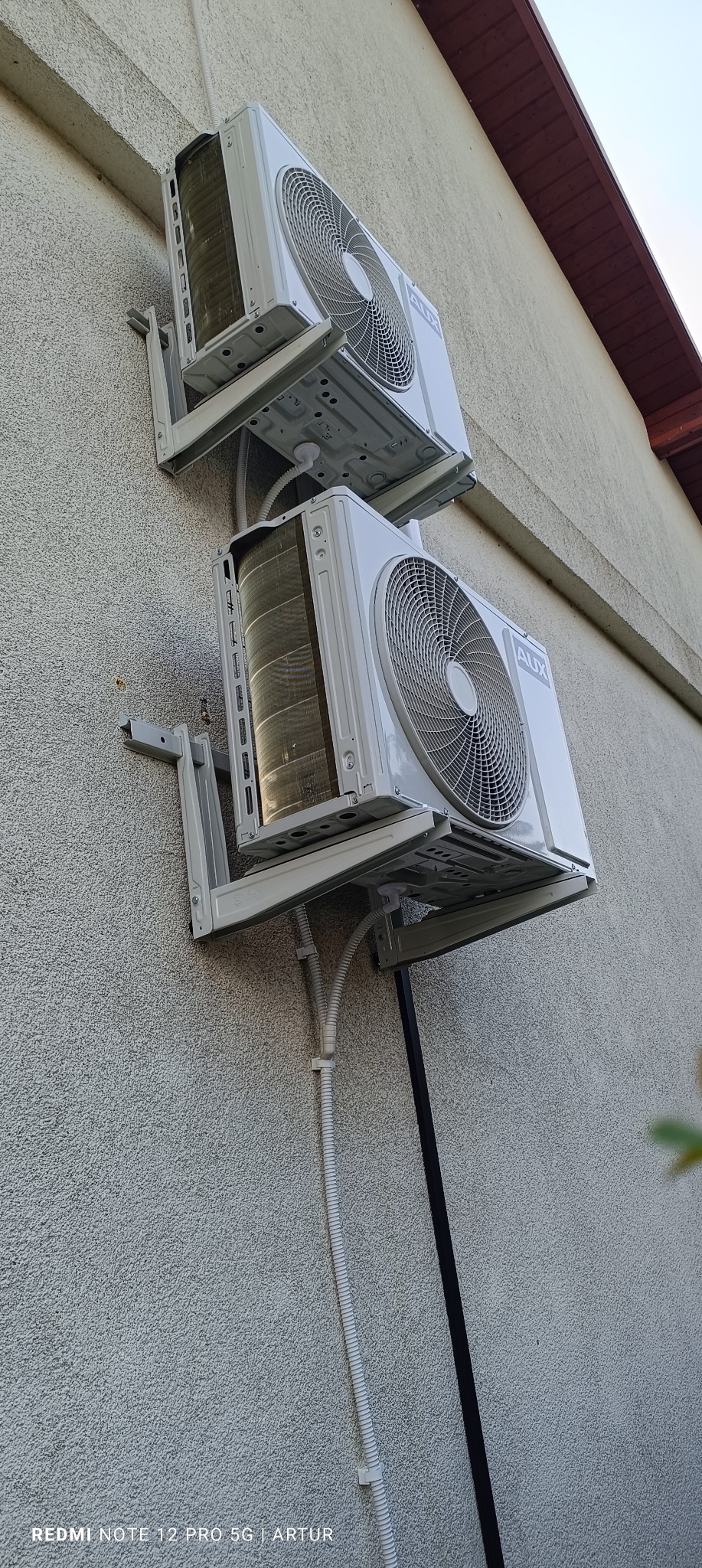 Dwa klimatyzatory marki AUX zamontowane na szarej, teksturowanej elewacji budynku z widocznymi przewodami instalacyjnymi.