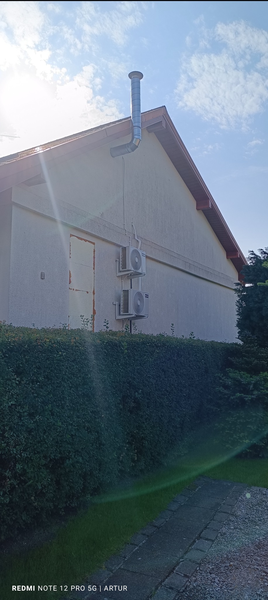 Dwa klimatyzatory marki AUX zamontowane na ścianie budynku obok zamurowanych drzwi, z widocznym kominem wentylacyjnym i zielonym żywopłotem na pierwszym planie.