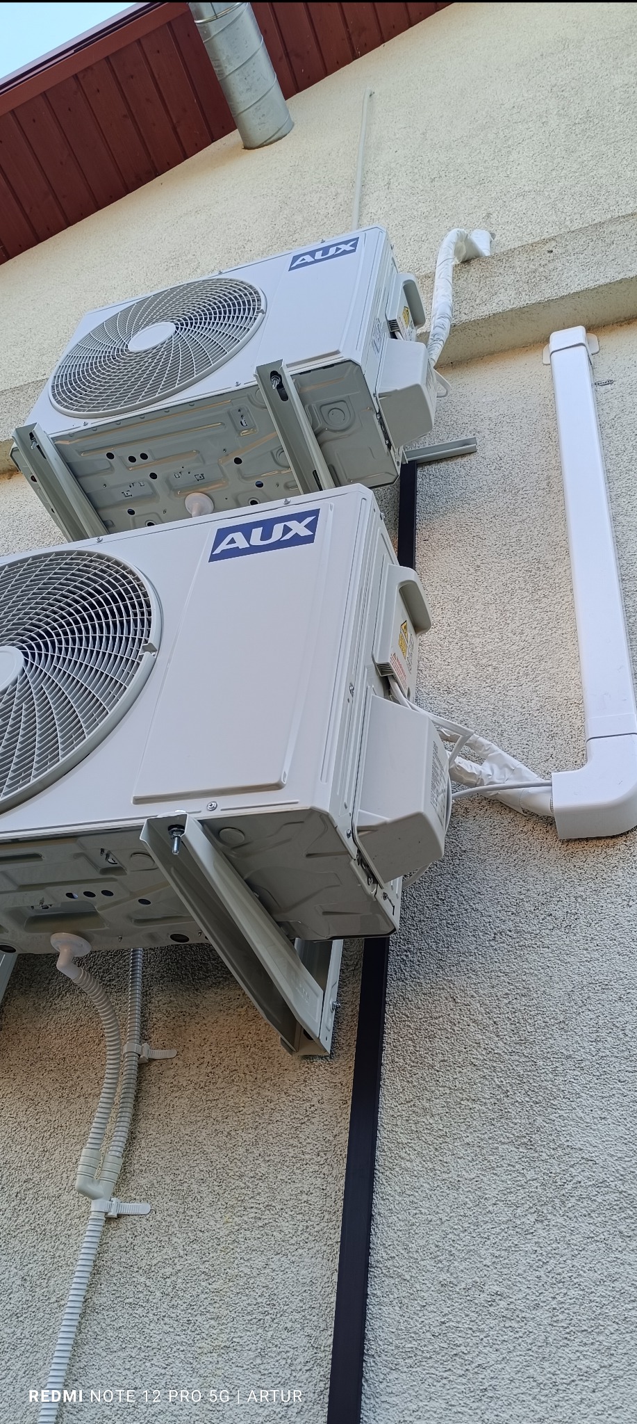 Dwa agregaty klimatyzacyjne marki AUX zamontowane jeden nad drugim na elewacji budynku, widoczne rury odprowadzające skropliny i przewody elektryczne.