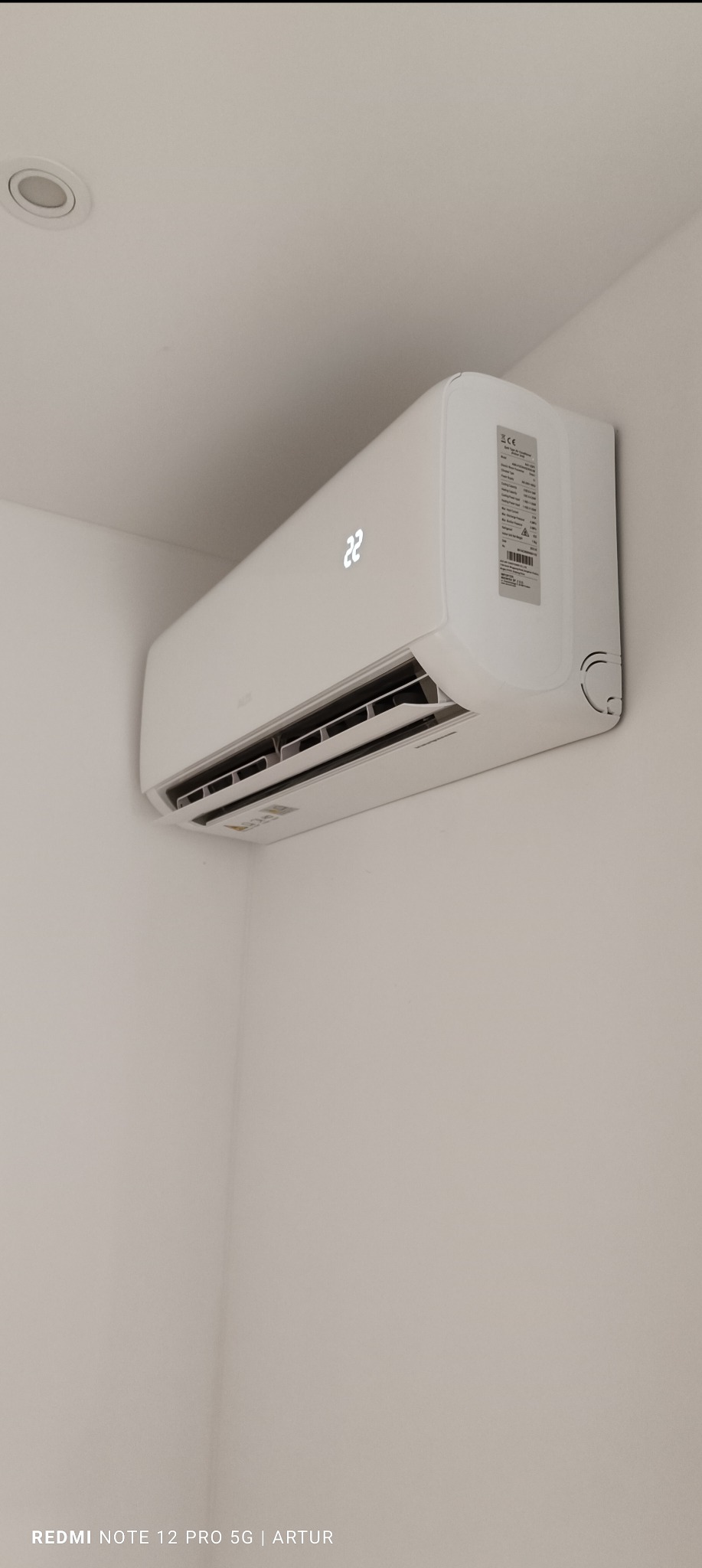 Biała jednostka klimatyzacji ściennej z wyświetlaczem temperatury 22 stopnie, zamontowana w rogu pomieszczenia o białych ścianach i suficie, widoczna z boku.