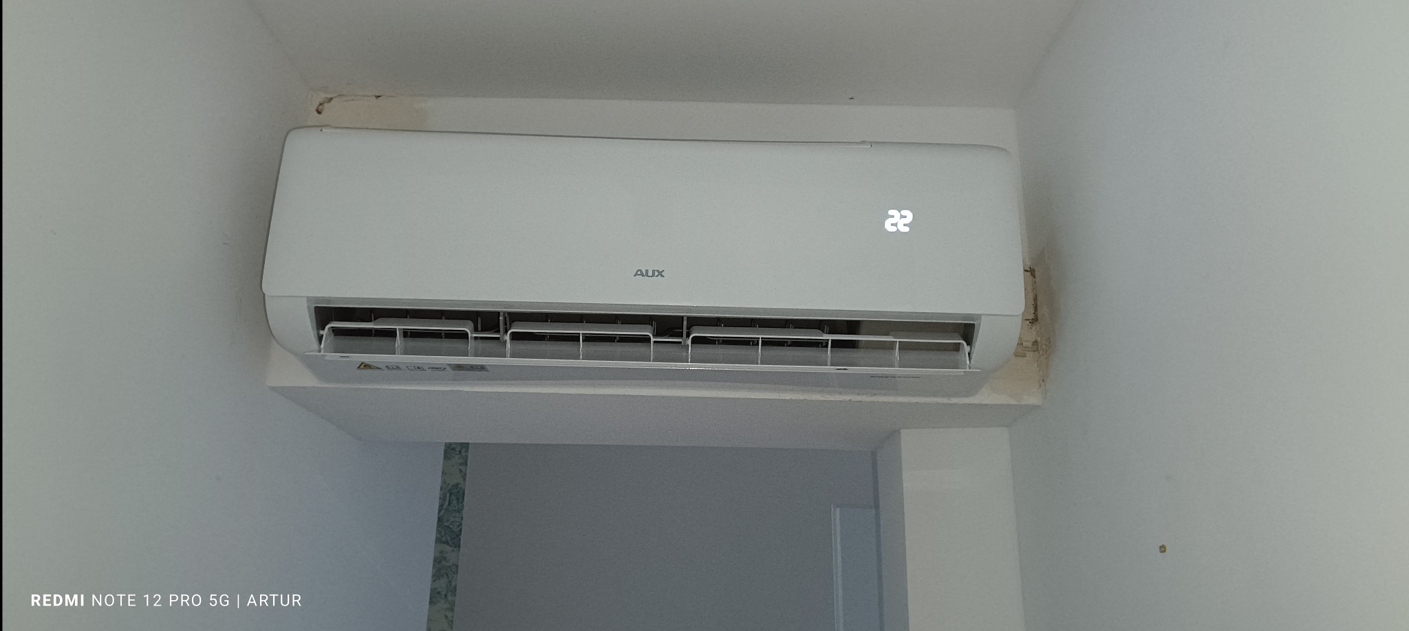 Biała klimatyzacja marki AUX zamontowana na ścianie, wyświetlacz pokazuje temperaturę 22 stopnie Celsjusza, widoczne niedoskonałości wykończenia ściany w rogu.