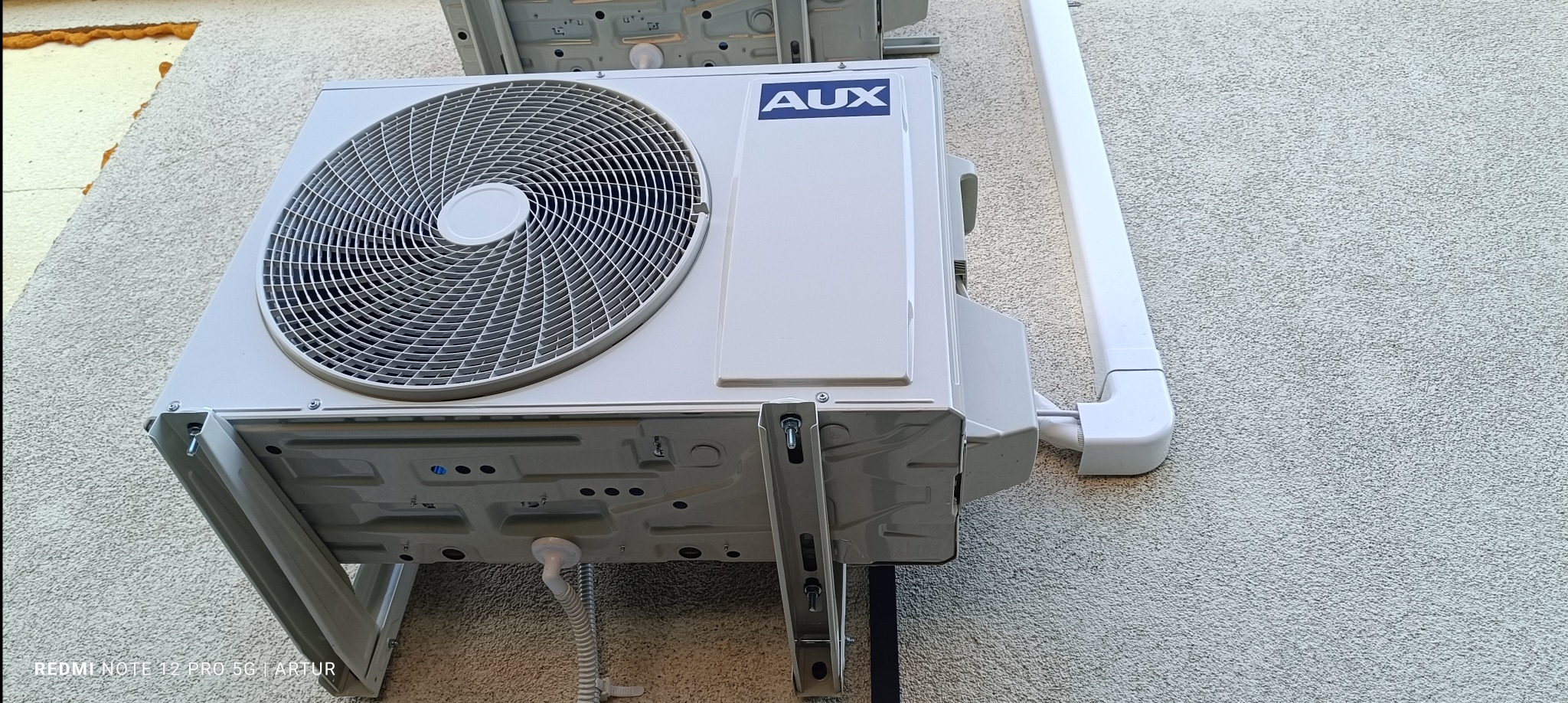 Zewnętrzna jednostka klimatyzacji AUX zamontowana na szarej elewacji budynku, widoczne rury odprowadzające skropliny i elementy mocujące.