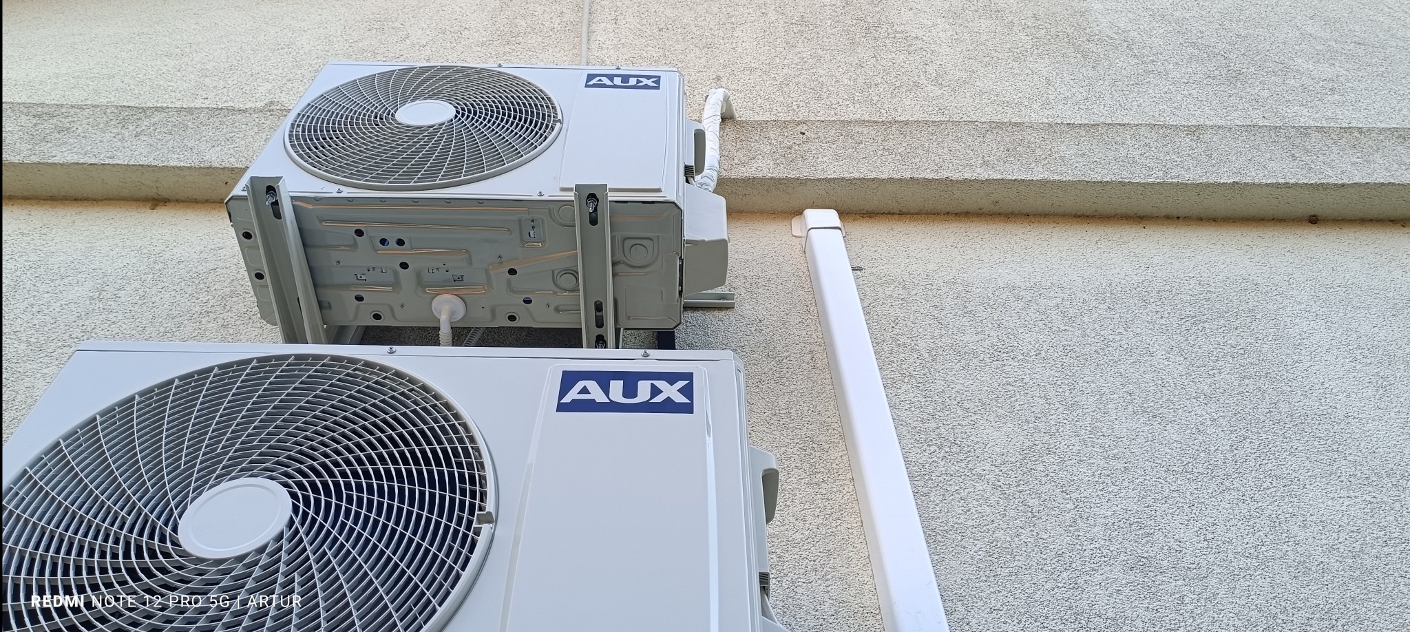 Dwa zamontowane na ścianie budynku klimatyzatory marki AUX, jeden nad drugim, z widocznymi rurami odprowadzającymi skropliny.