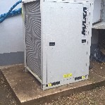 Szara pompa ciepła marki Nulxa na betonowej podstawie przy ścianie budynku, widoczny niebieski wąż. Minimalistyczny design, prosta bryła.