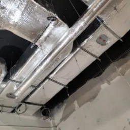 Izolowane kanały wentylacyjne i rury pod sufitem, zamocowane na metalowych szynach, widoczne przewody i fragment surowej ściany.