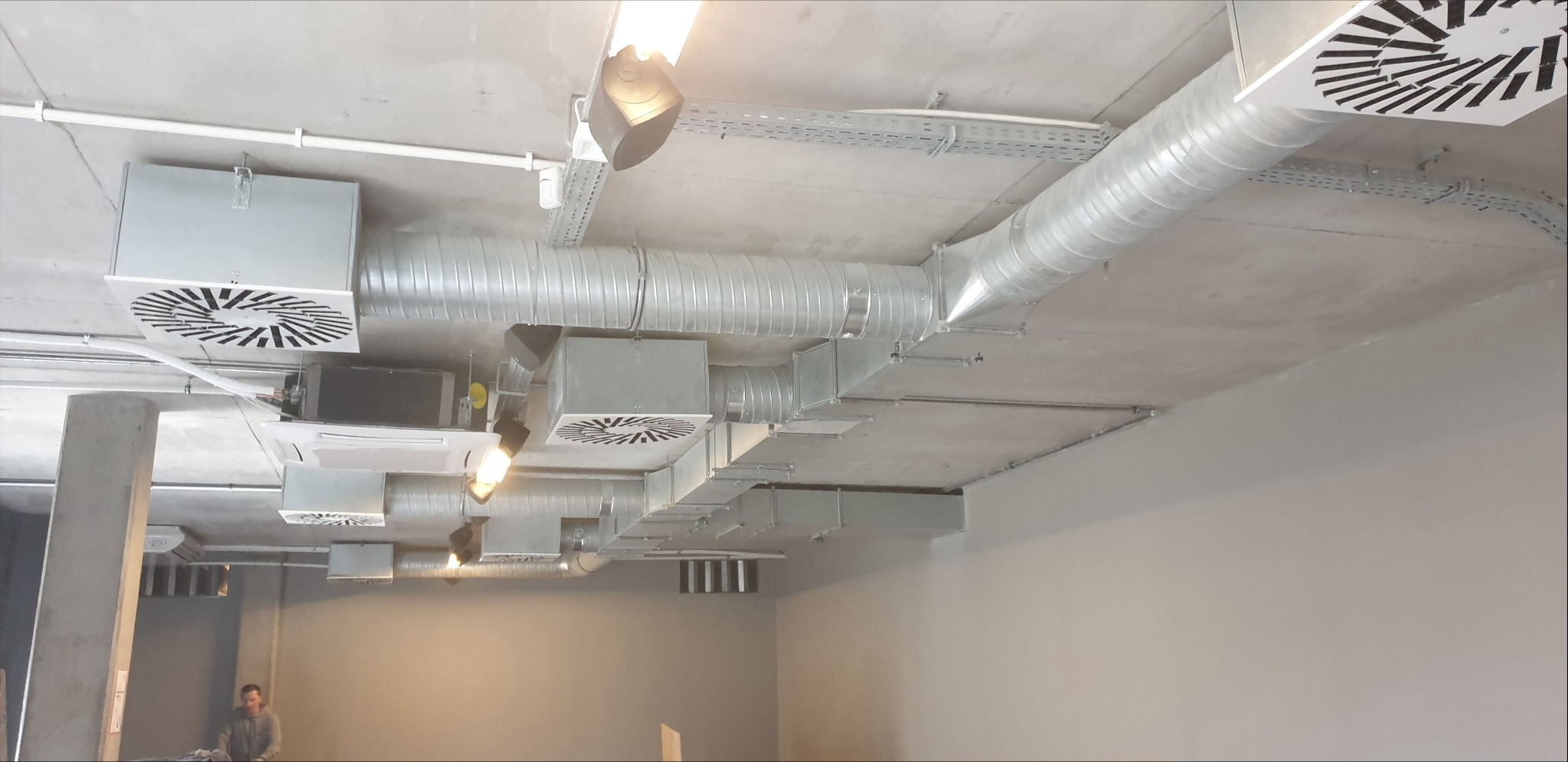 Kompleksowy system wentylacji i klimatyzacji w biurze, widoczne kanały wentylacyjne, klimatyzatory i kratki wentylacyjne na suficie z instalacją elektryczną.