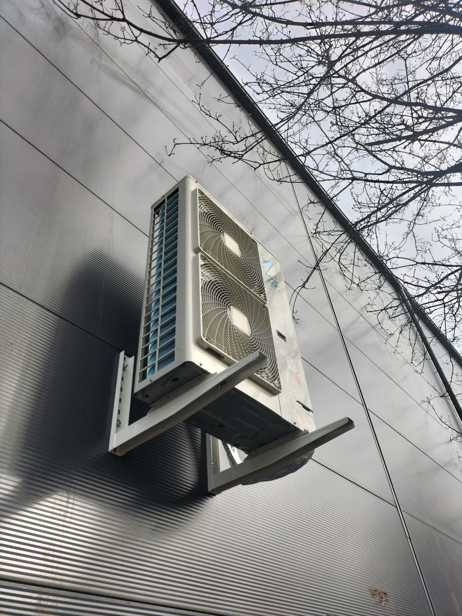 Zewnętrzna jednostka klimatyzacyjna zamontowana na metalowej elewacji budynku, widoczne drzewo bez liści w tle.