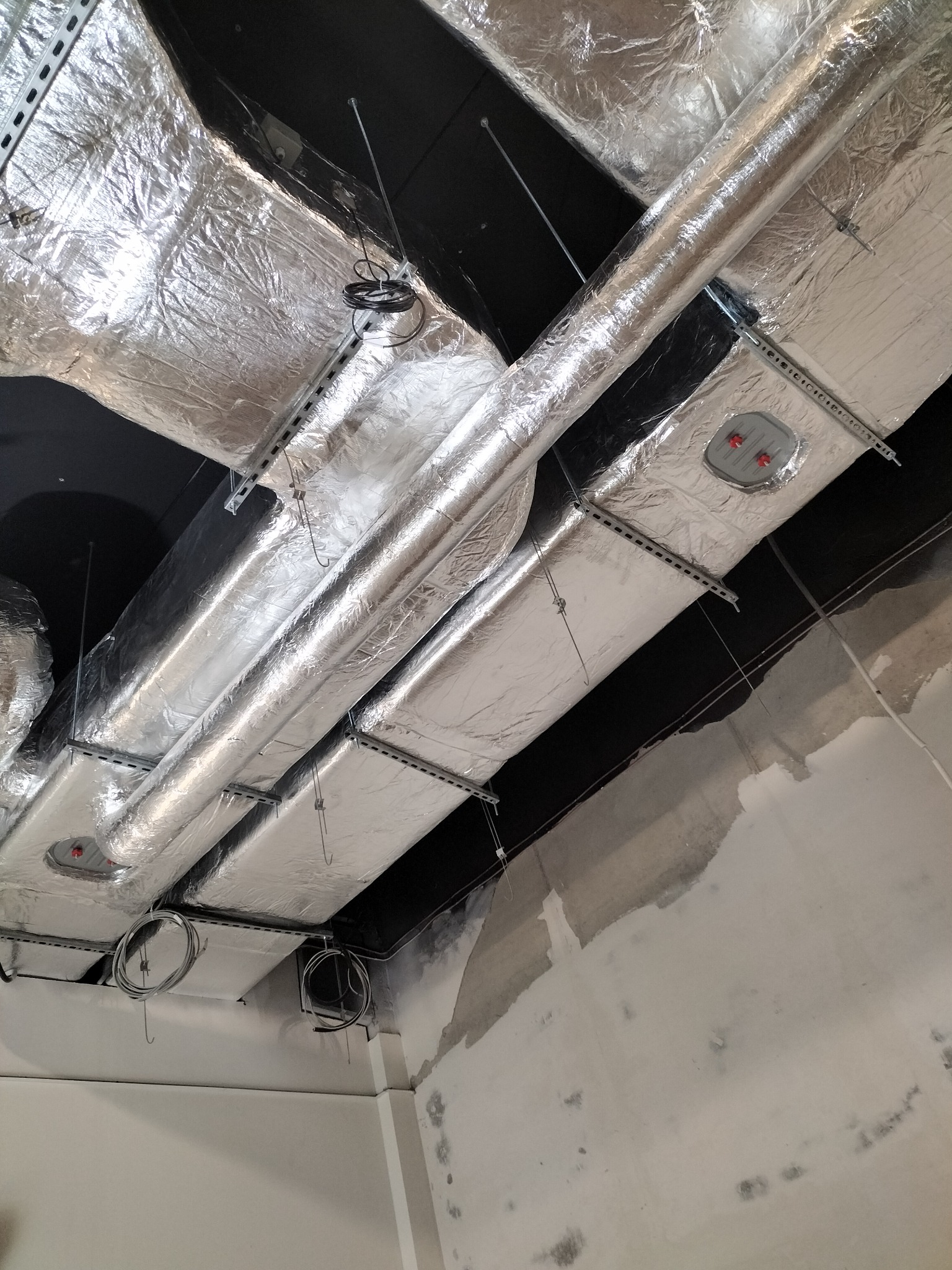 Izolowane kanały wentylacyjne i rury pod sufitem, zamocowane na metalowych szynach, widoczne przewody i fragment surowej ściany.