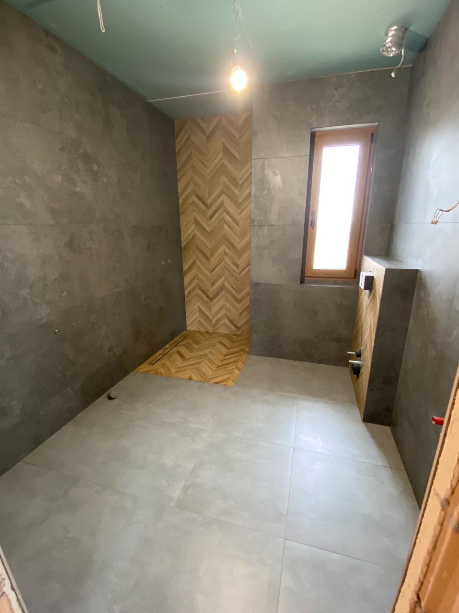 Łazienka w trakcie remontu: szare płytki podłogowe i ścienne, drewniana mozaika ułożona w jodełkę na ścianie prysznica i obudowie instalacji, surowy stan wykończenia, widoczne instalacje...