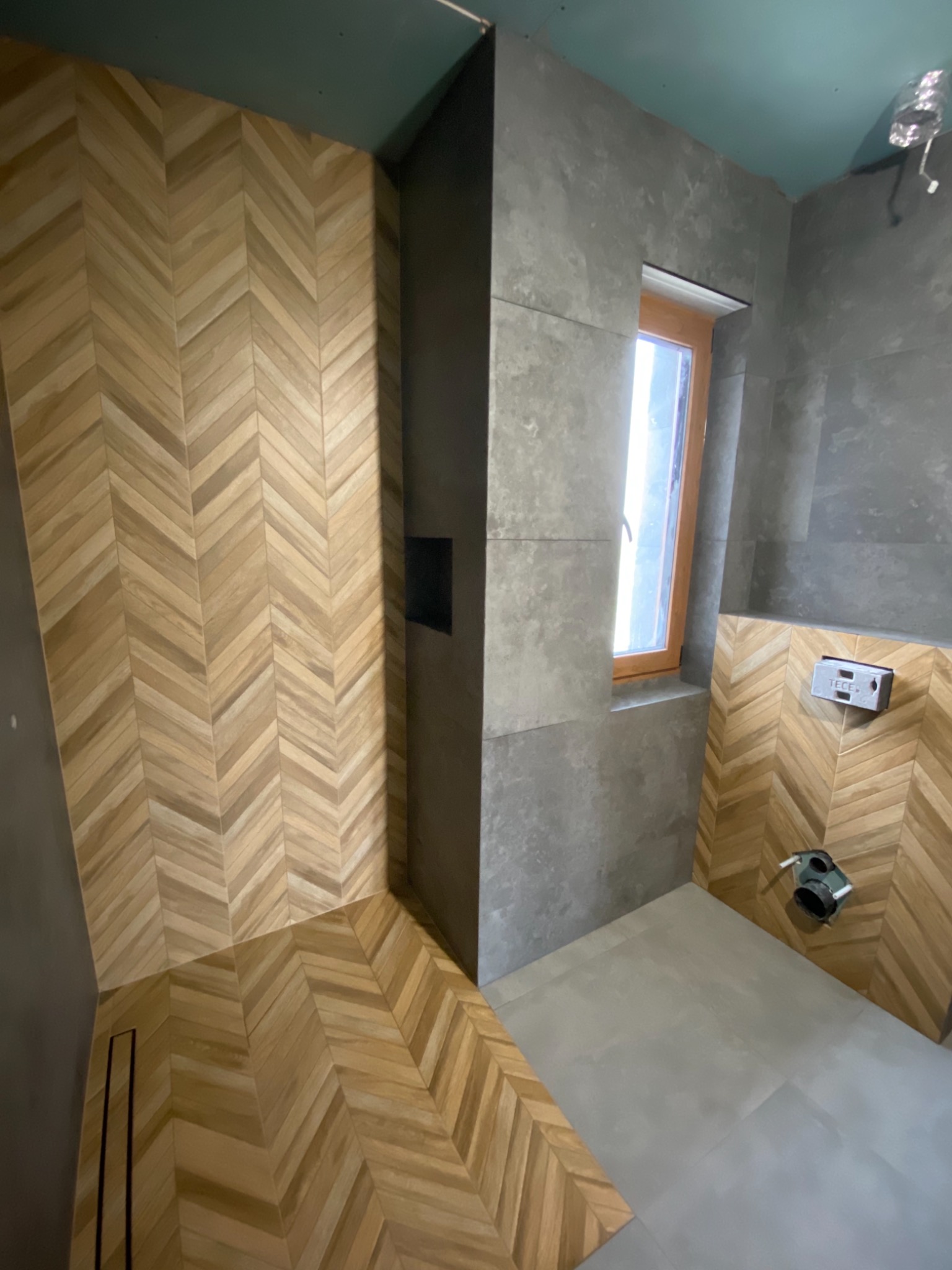 Łazienka w trakcie wykańczania: drewnopodobne płytki ułożone w jodełkę na ścianie i podłodze prysznica, szare płytki na pozostałych ścianach, okno z drewnianą ramą i widoczne elementy instalacji.